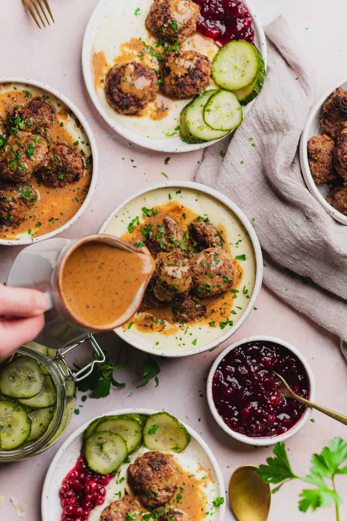 hand pouring brown gravy over top of keto swedish meatballs, with lingonberry sauce and pickled cucumbers nearby. 