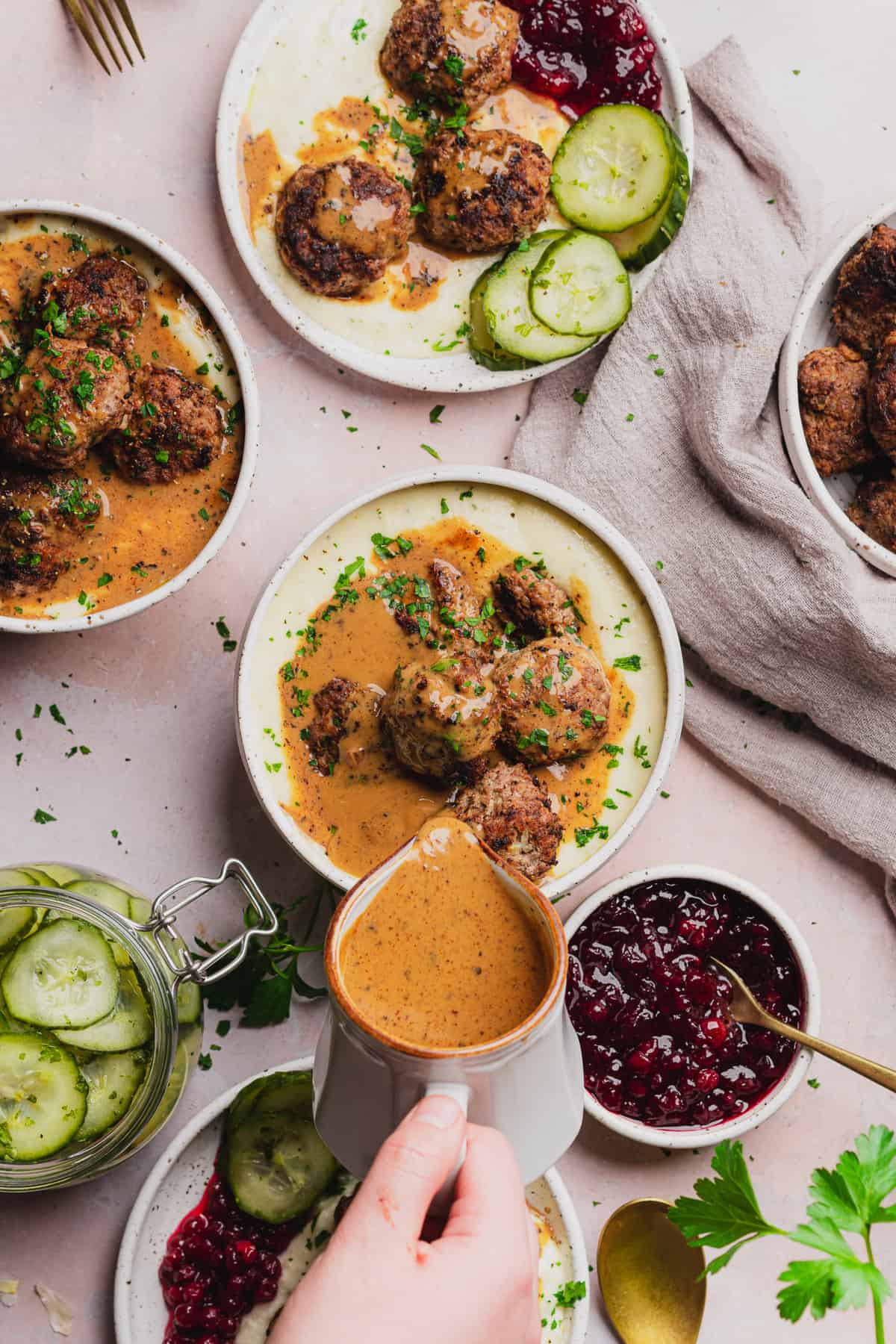hand pouring brown gravy over top of swedish meatballs with cucumber salad and lingonberry sauce