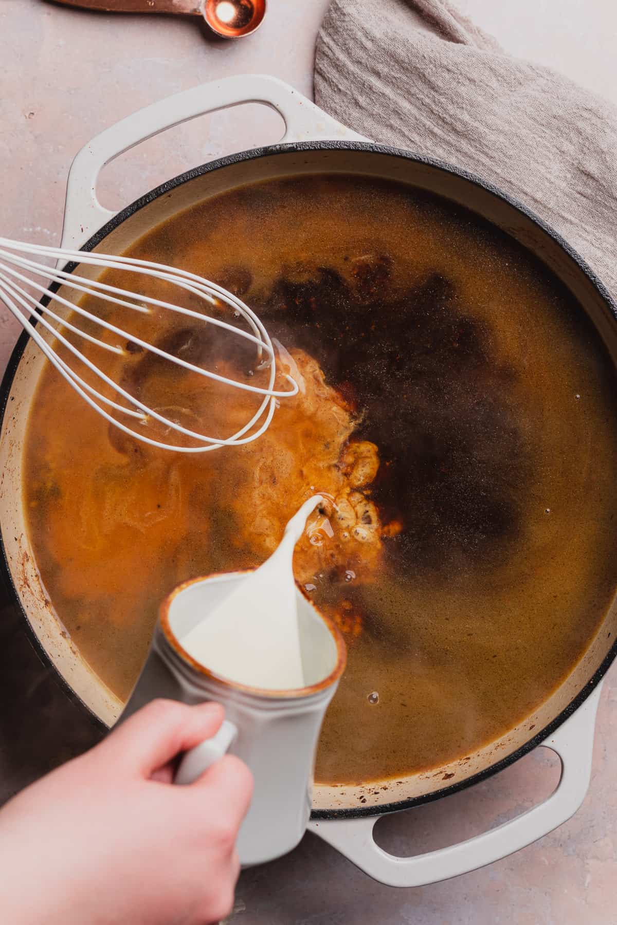 hand pouring cream into a skillet with broth and whisking simultaneously. 