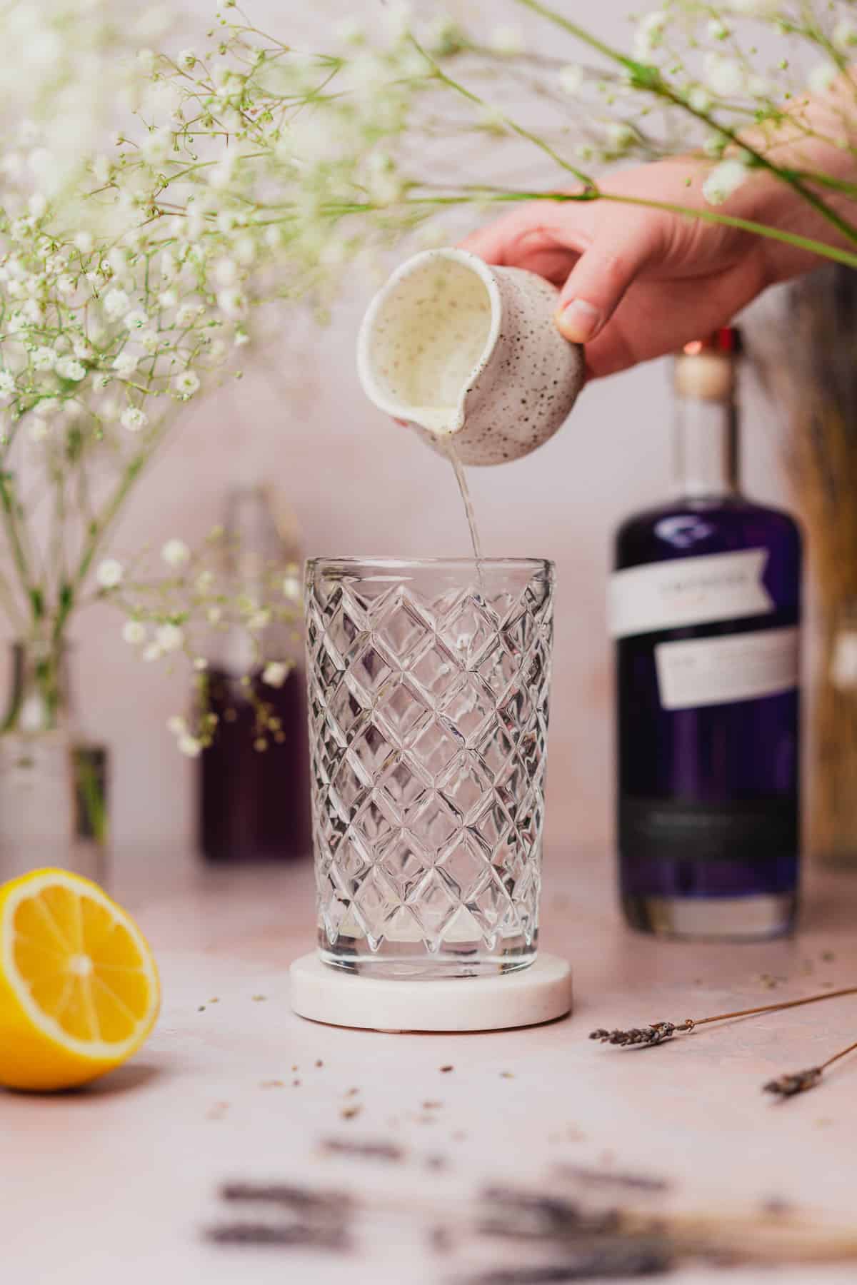 hand pouring lemon juice into a glass cocktail shaker with empress gin and baby's breath flowers surrounding it 