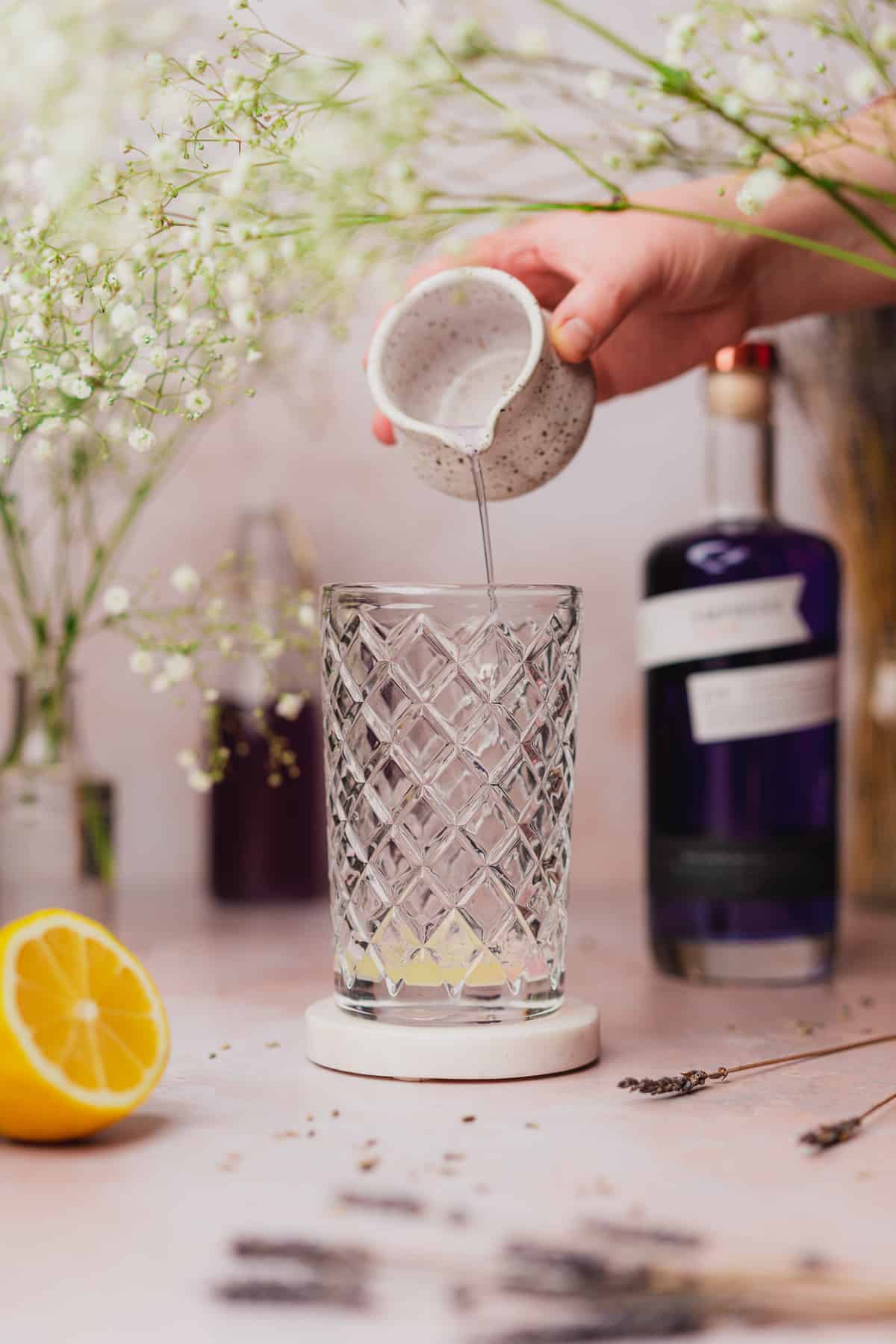 hand pouring simple syrup into a glass cocktail shaker surrounded by lemon, empress gin and baby's breath flowers 