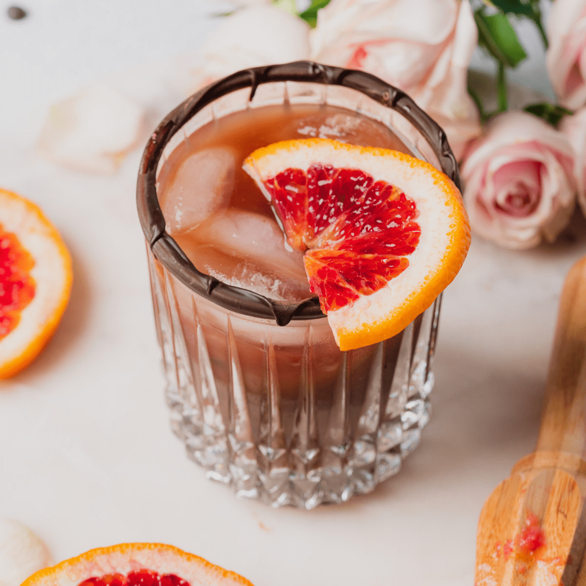 Chocolate Old Fashioned with Blood Orange surrounded by lovely light pink spray roses, a citrus reamer, and orange slices