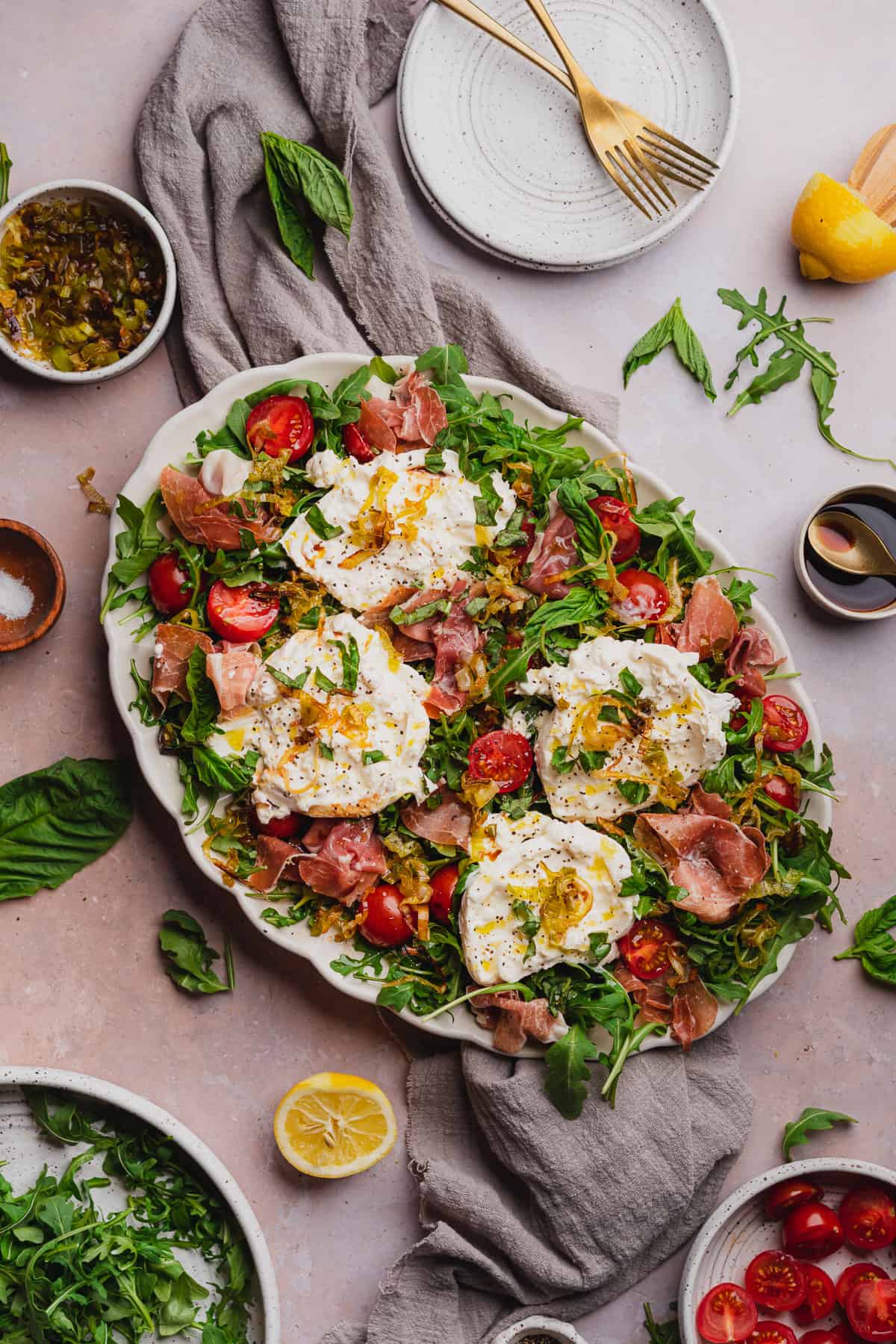 flat lay shot of a burrata and arugula salad with prosciutto, tomatoes, basil, lemon, fried leeks, and balsamic vinegar on a white scalloped plate. 