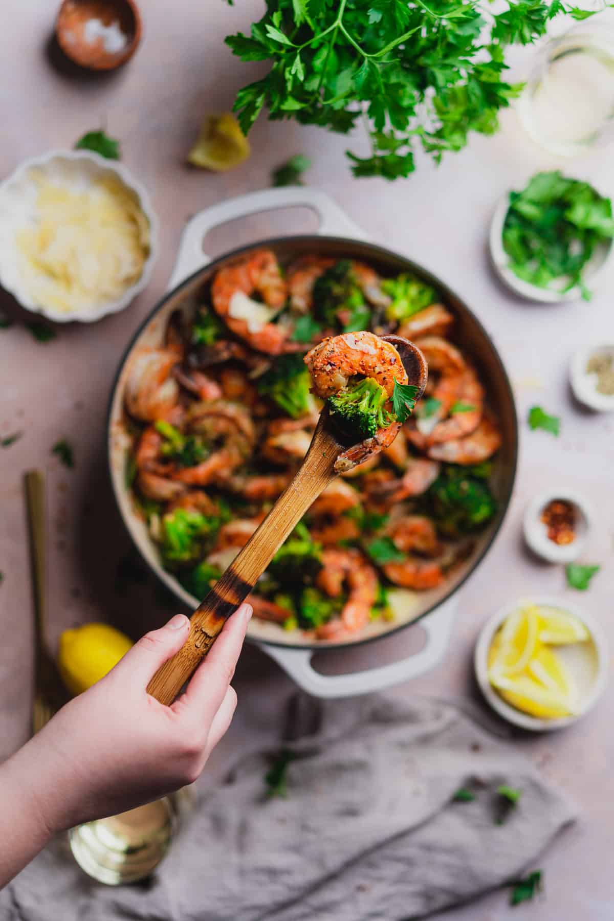 hand holding a wooden spoonful of keto shrimp and broccoli over a skillet. 