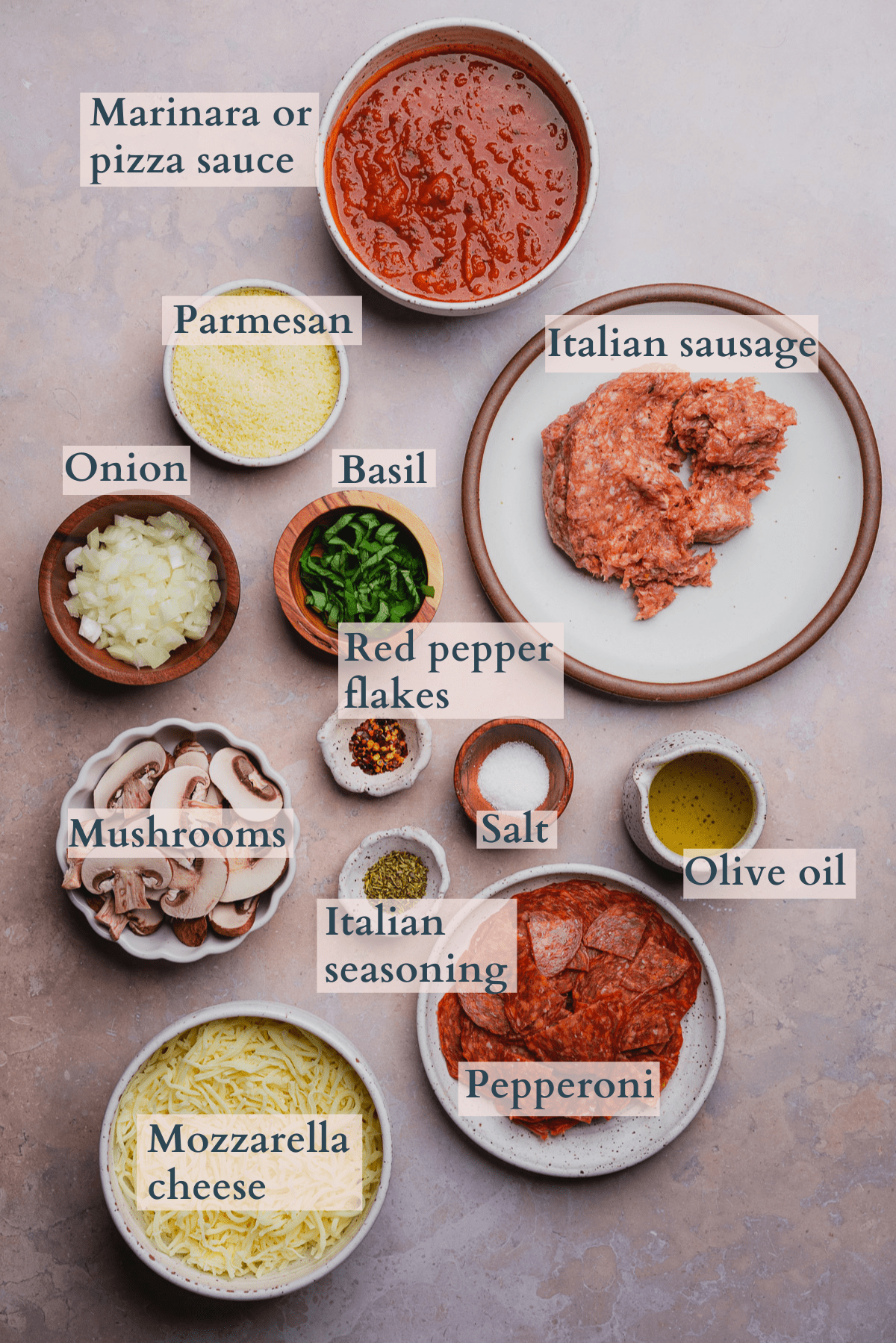 pizza bowl recipe ingredients with text to denote different ingredients, featuring marinara sauce, basil, mozzarella, parmesan, Italian sausage, pepperoni, red pepper flakes, Italian seasoning, salt, mushrooms, onion, and olive oil. 
