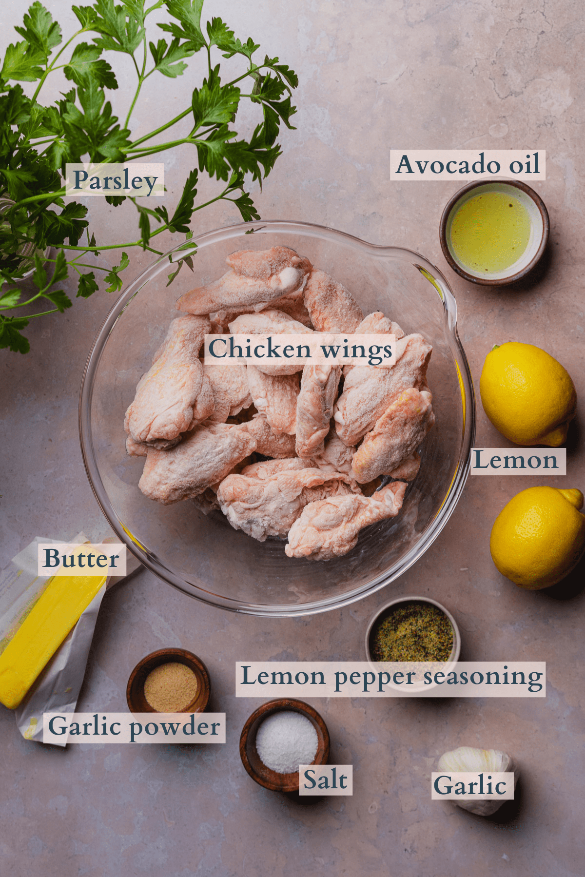 ingredients to make air fryer lemon pepper wings, with raw chicken wings, lemons, avocado oil, garlic, garlic powder, lemon pepper seasoning, salt, butter, and parsley.