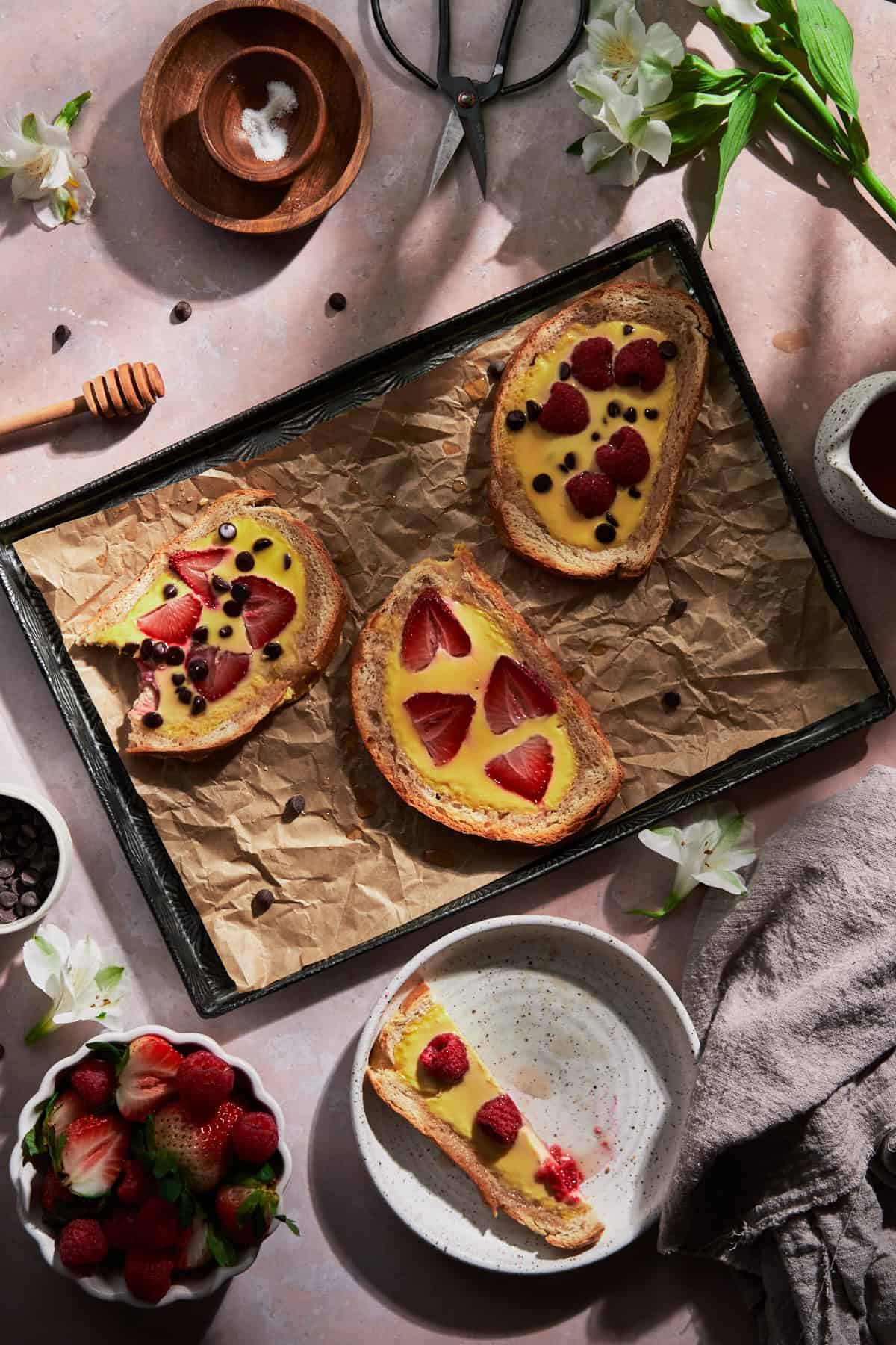 flat lay scene of custard yogurt toast with berries, maple syrup, fruit, chocolate chips, and flowers. 