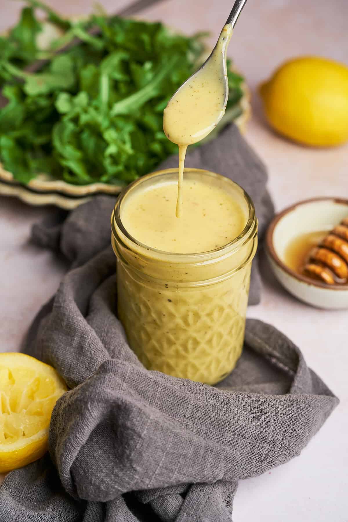 jar full of honey lemon vinaigrette salad dressing, with fresh lemon and honey in the background, along with a simple green salad and a spoonful of dressing falling off into the jar. 