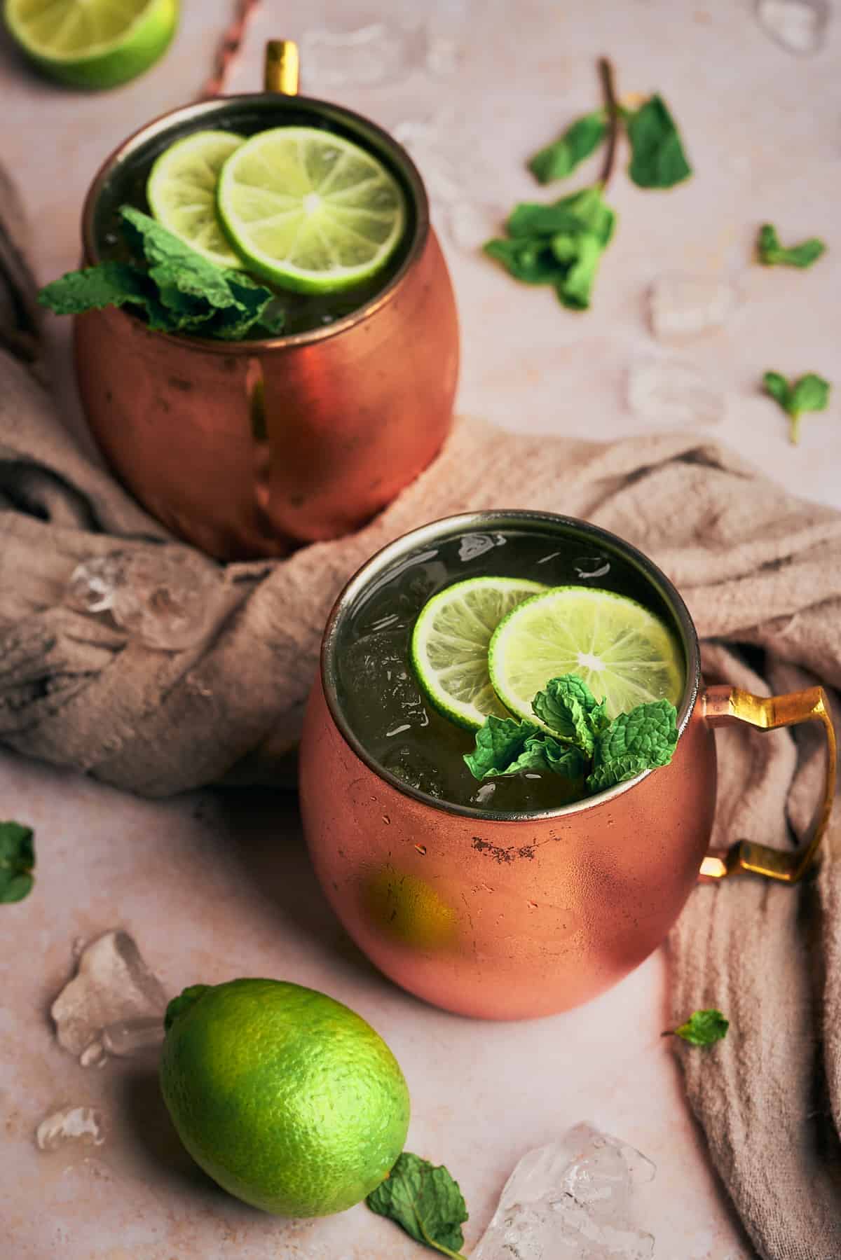 two Kentucky mules in copper mugs with fresh mint and lime slices, a grey linen towel between them, with lime slices, mint leaves and ice on the scene. 