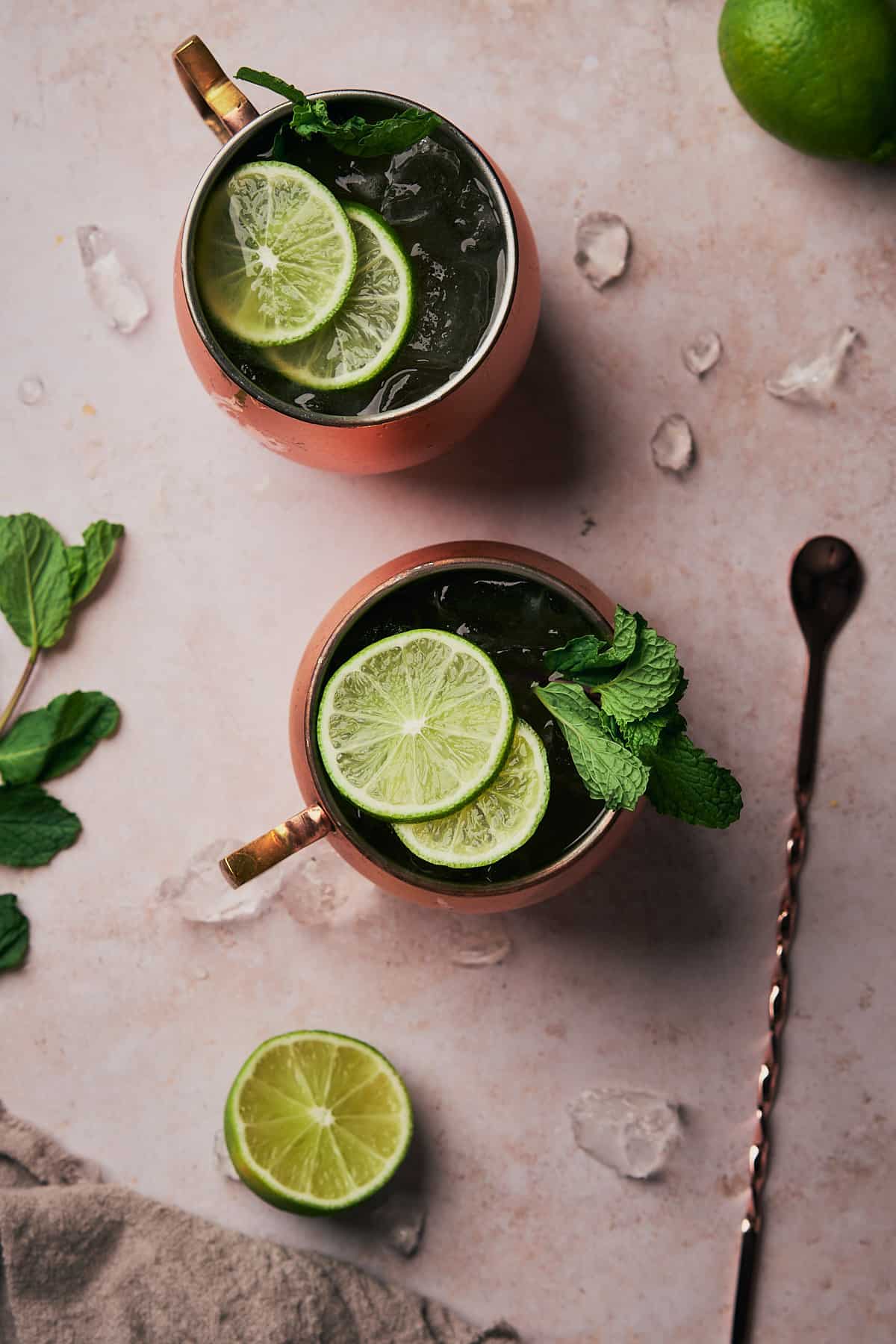 a flat lay scene of two kentucky mules in copper mugs, with ice, lime slices, fresh mint, and a bar spoon on the scene. 