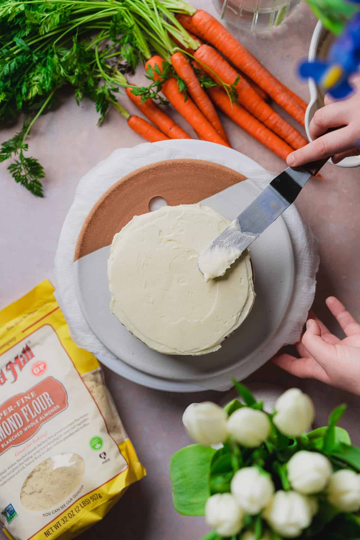 hand spreading cream cheese frosting with an angled spatula on a keto carrot cake, with fresh white tulips, fresh carrots, and a bag of bob's red mill almond flour nearby. 