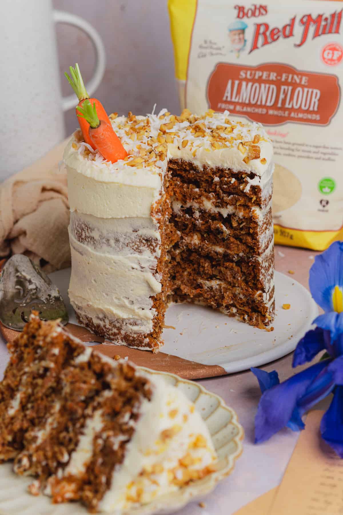 tall keto carrot cake recipe with a large slice taken out, and bob's red mill almond flour in the back, with a slice in front.