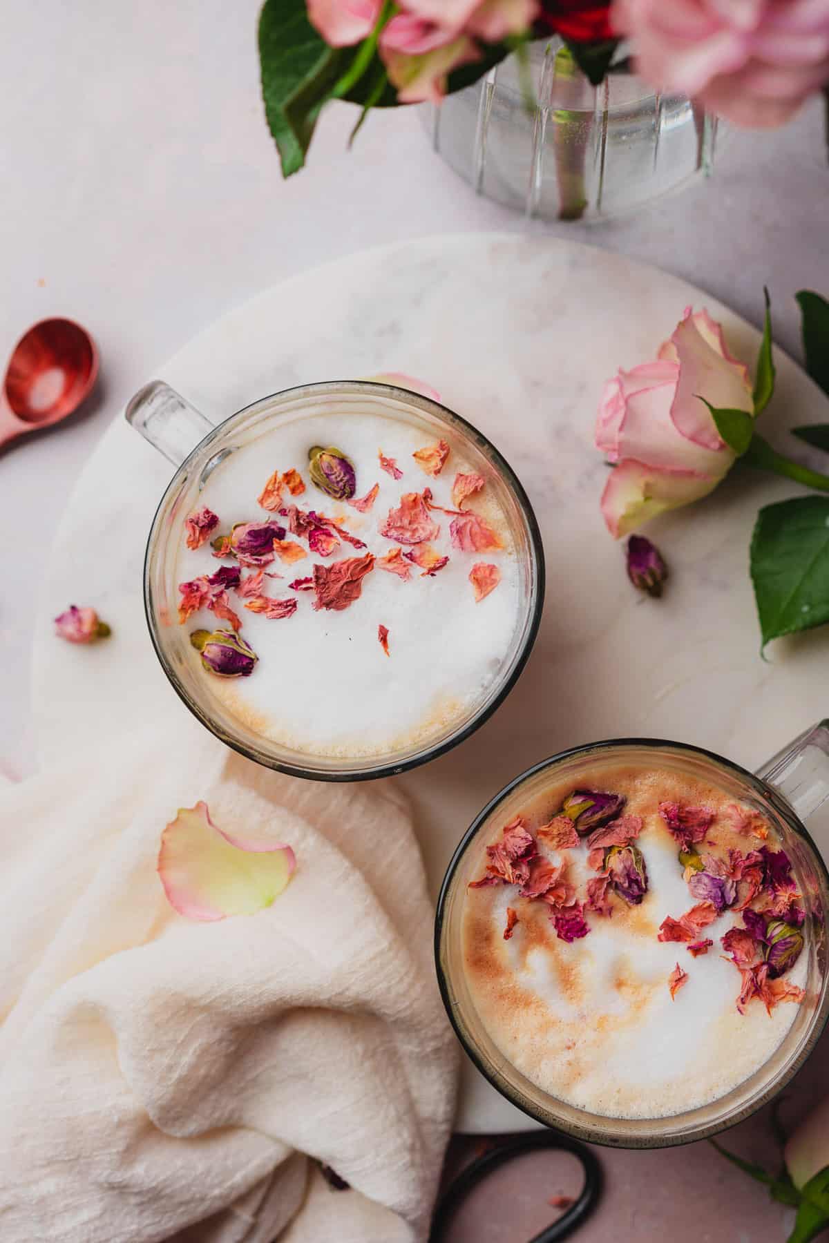 two mugs of a rose latte topped with rose petals and buds, and surrounded by fresh roses. 