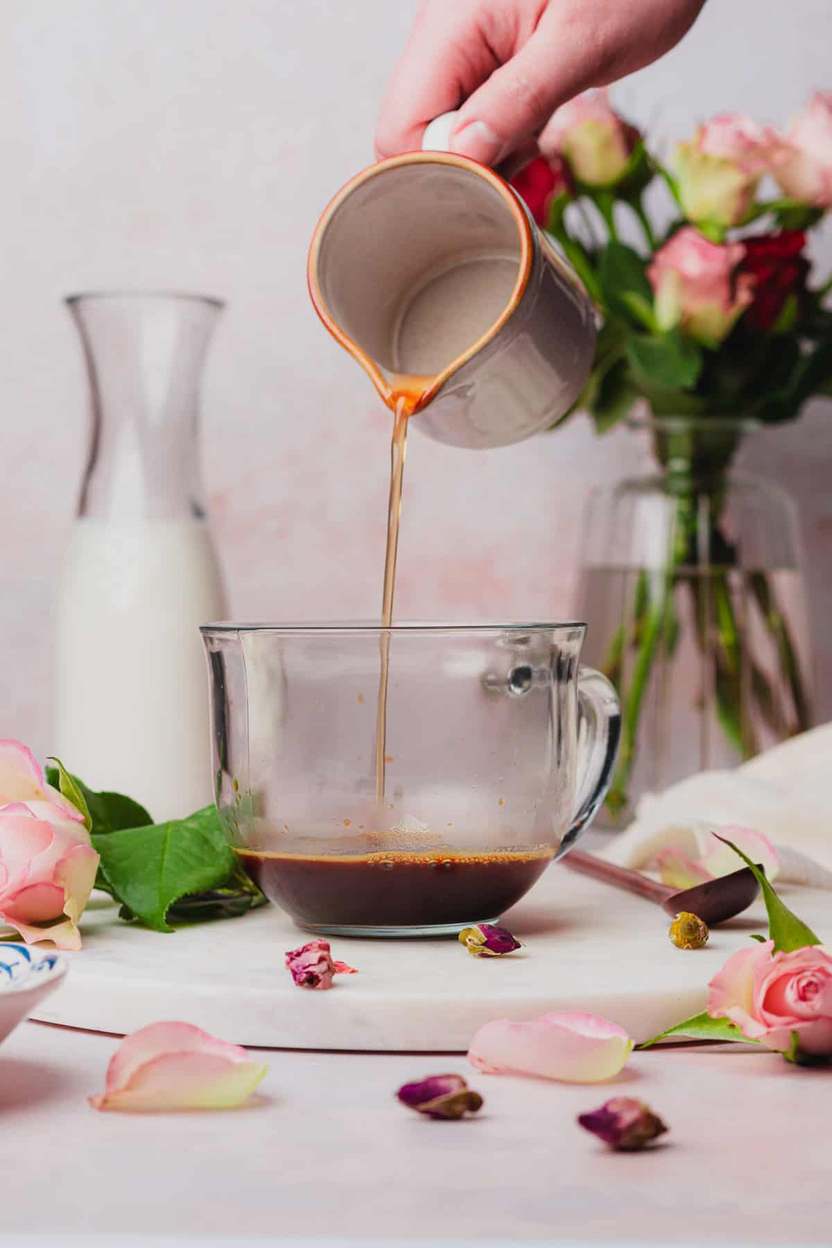 hand pouring in rose simple syrup into a coffee mug with espresso in it, surrounded by milk and flowers. 