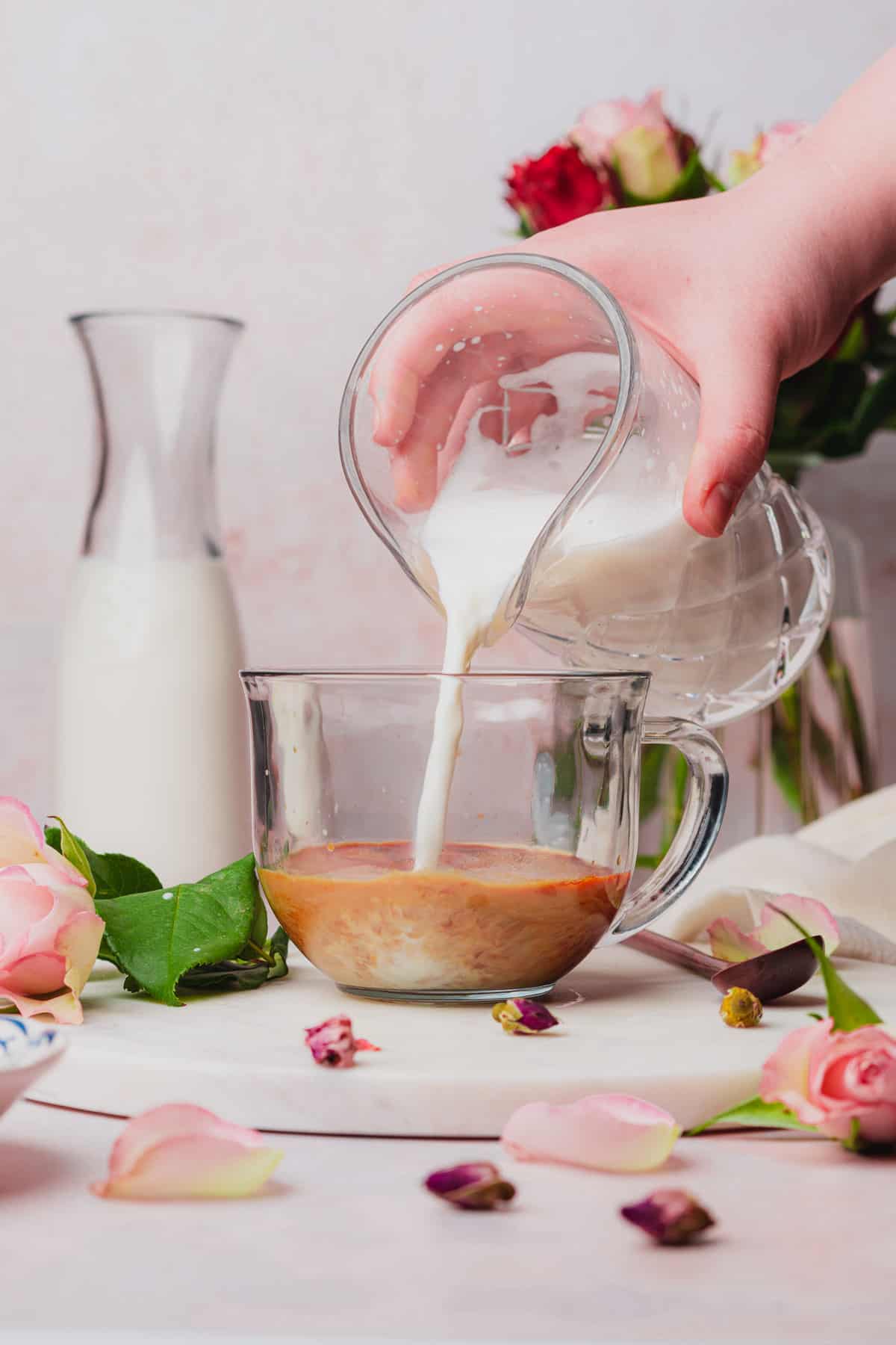hand pouring steamed milk into a mug surrounded rose petals, rose buds, and milk. 