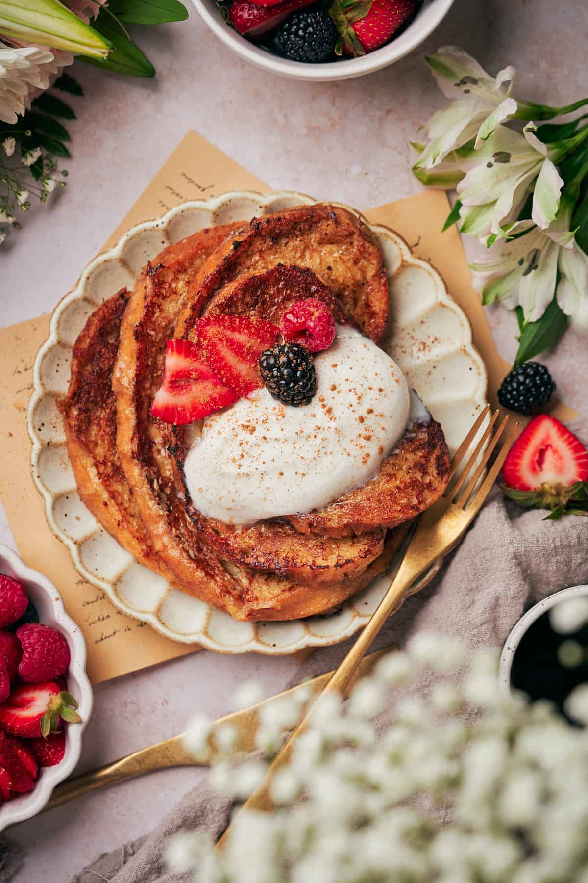Sourdough french toast with strawberries and blackberries, with a gold fork on a plate with flowers in the background.