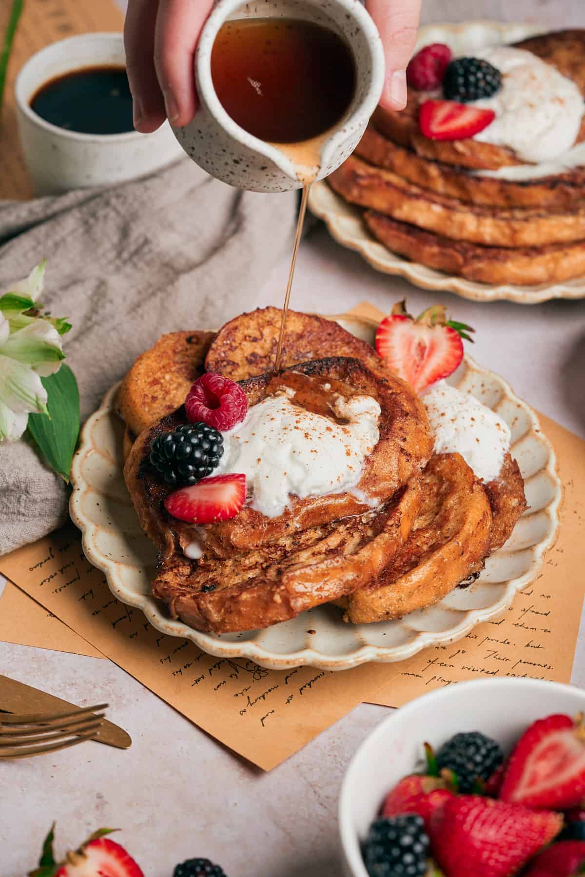 45 degree shot of a a stack of sourdough french toast with fresh berries and whipped cream on it. There is a hand pouring on maple syrup.