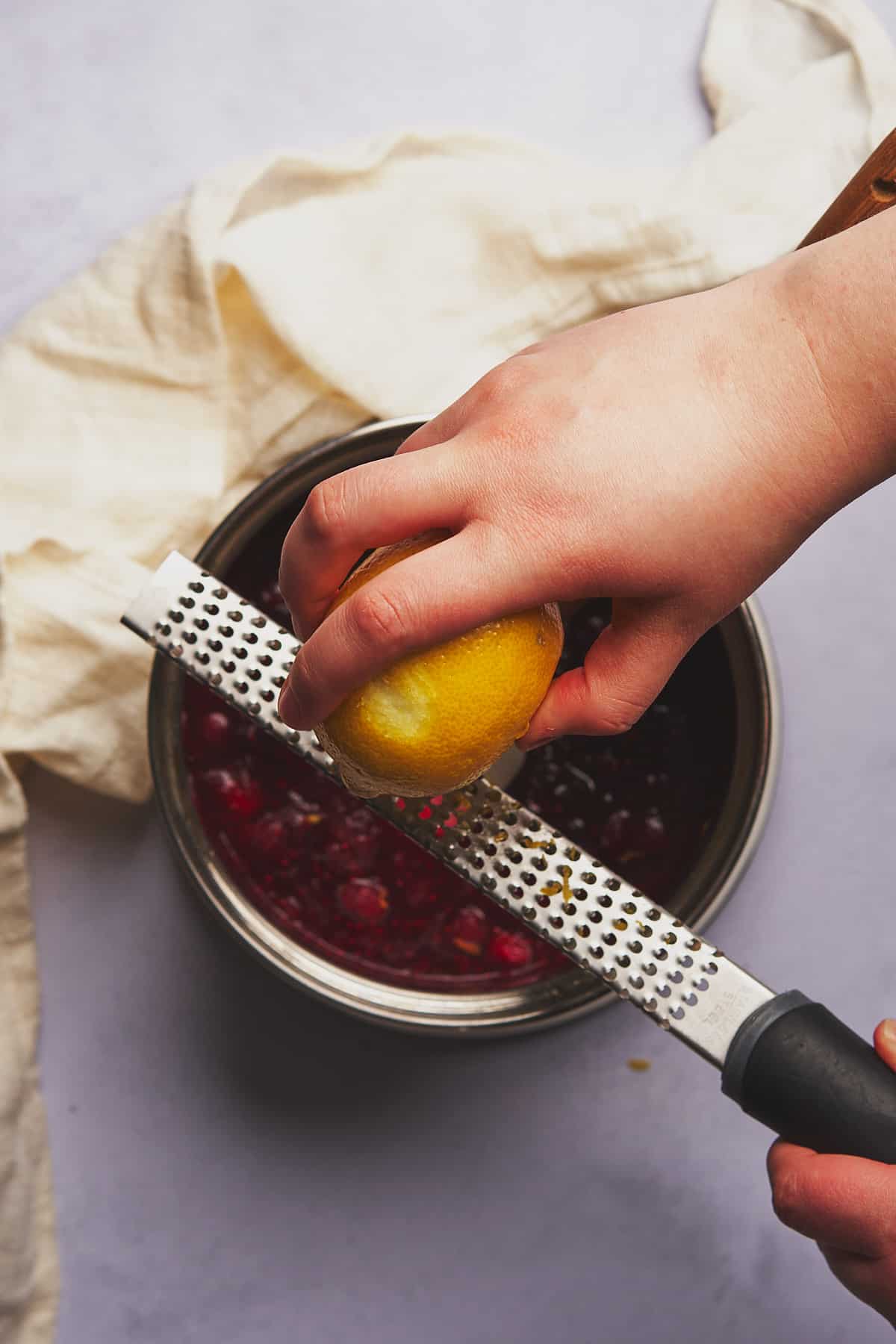 hand zesting a lemon against a zester into a pot of cranberry sauce. 