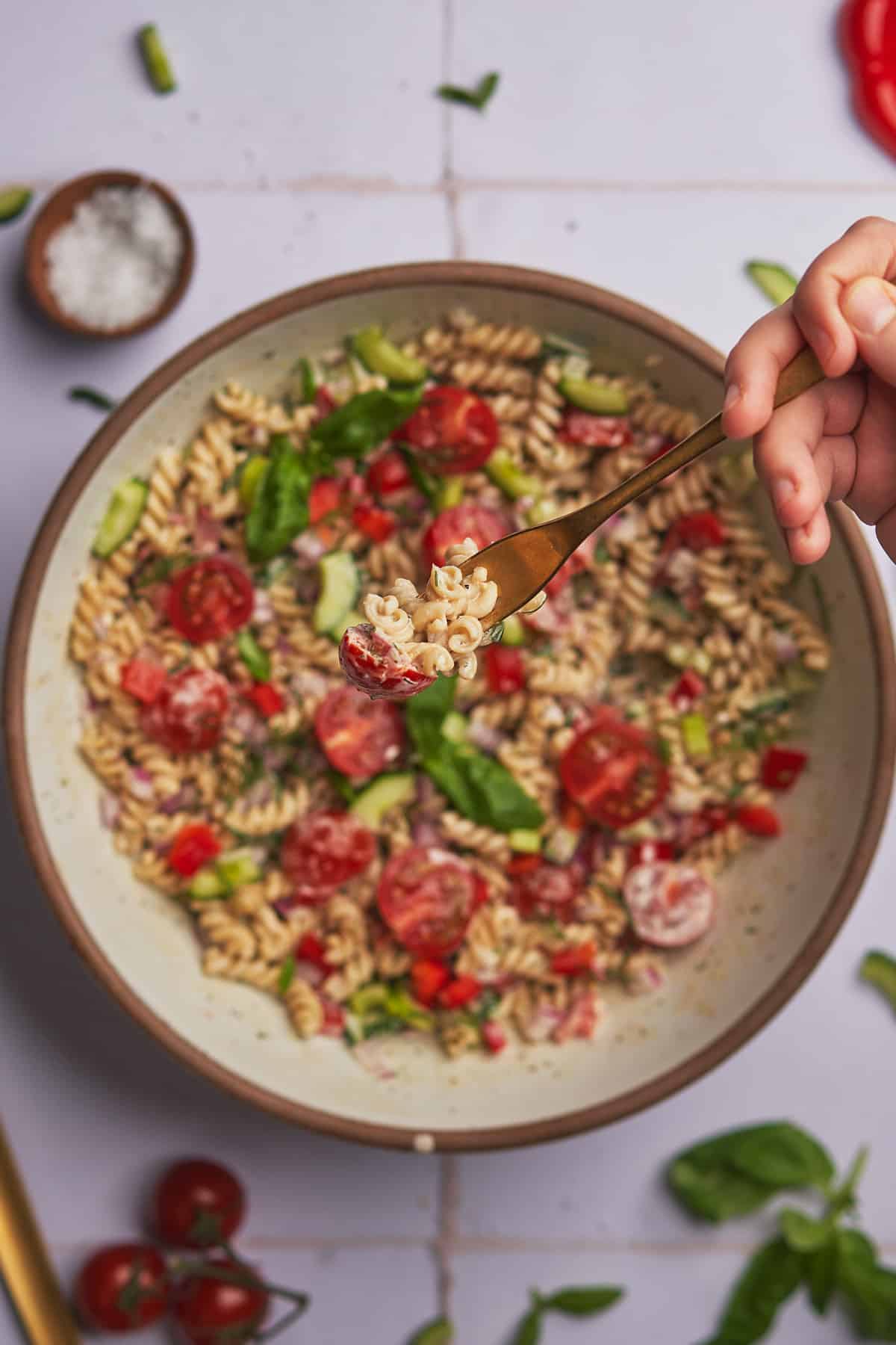 hand reaching into a large bowl of pasta salad with fresh vegetables and herbs.