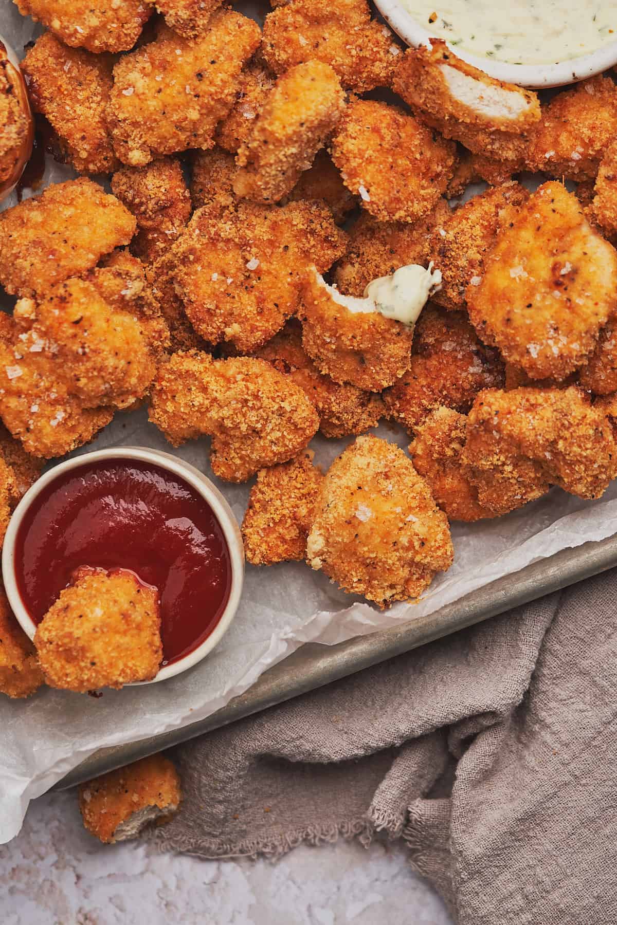 tray of keto chicken nuggets served with ranch, bbq sauce, and ketchup on a tray lined with white parchment paper, with one nugget dunked in ketchup. 