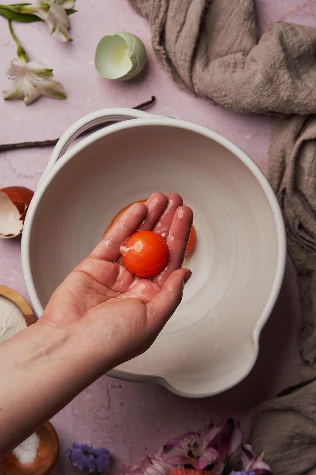 hand holding a bright orange egg yolk over a white ceramic bowl. 