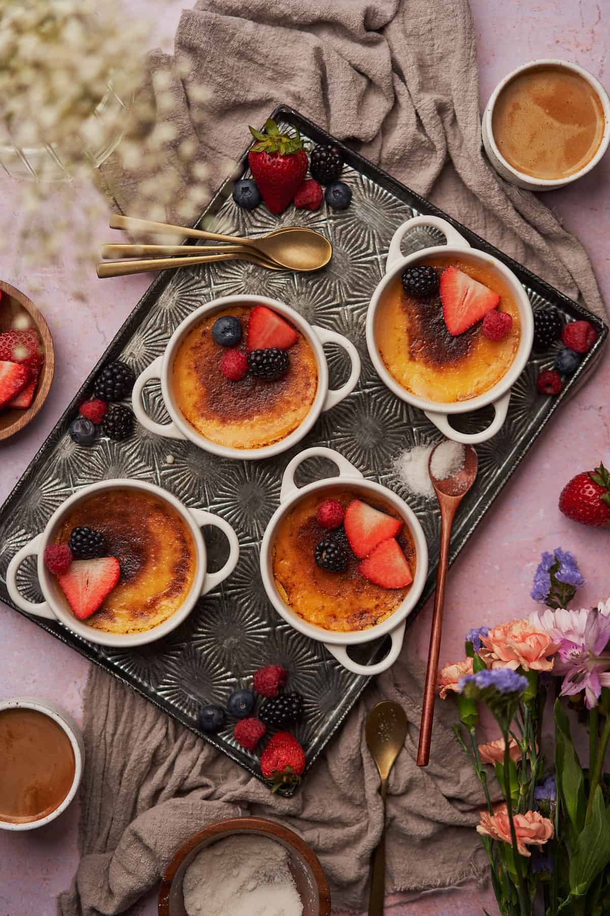 4 creme brulees in mini ramekins topped with berries, and flowers, and coffee surrounding the scene.  