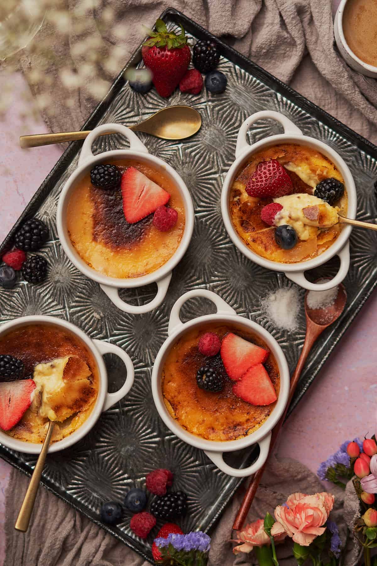 close up shot of 4 creme brulees topped with berries, and two of the ramekins have spoons that have broken the caramelized sweetener top to show of the egg custard in the middle.