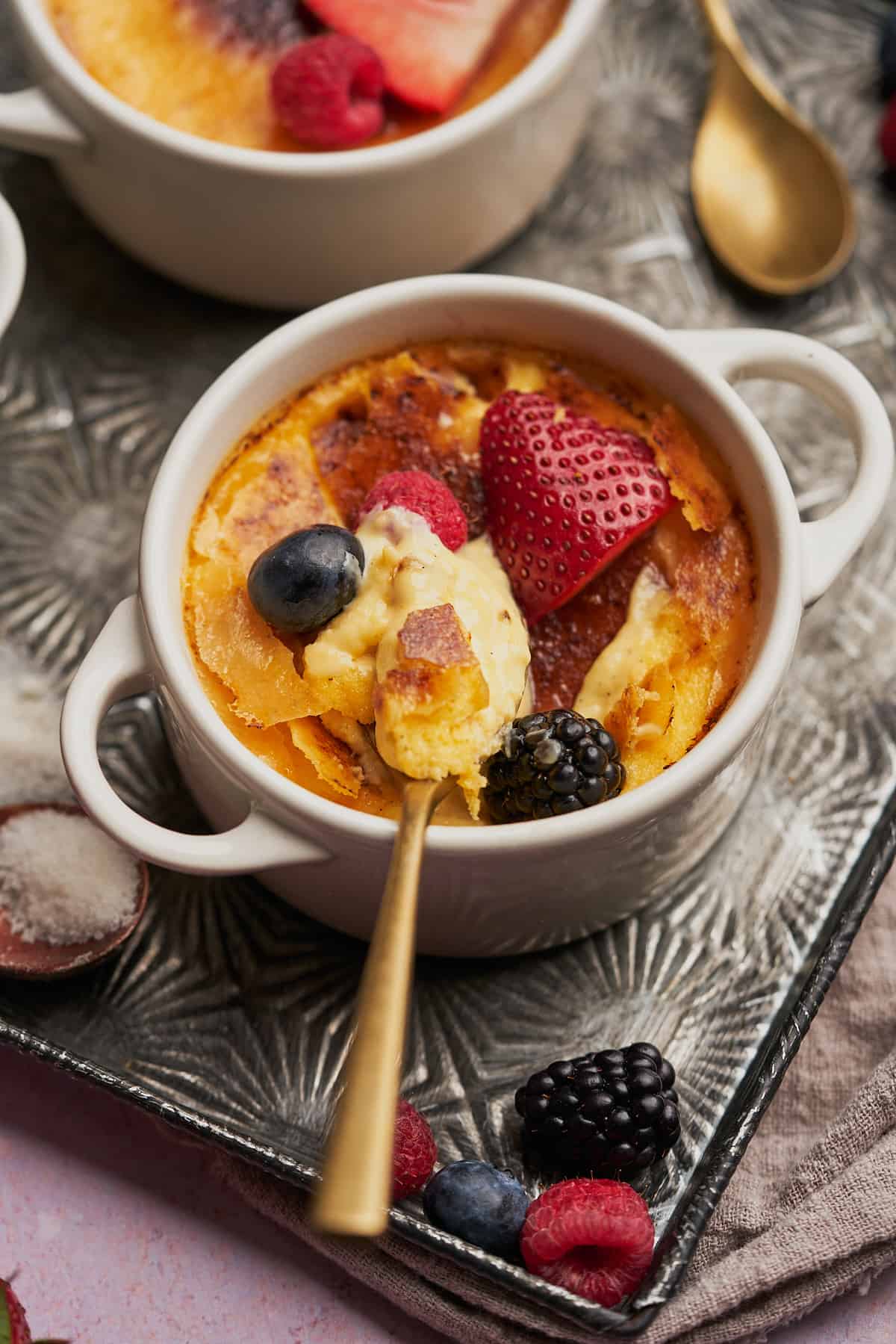 close up shot of keto creme brulee with strawberries, a raspberry, and a blackberry on top, with a spoon showing the inside of the custard.