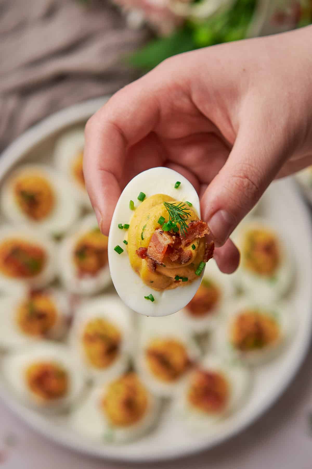 hand holding a keto deviled egg to the screen topped with chives, dill, bacon, and prosciutto. 