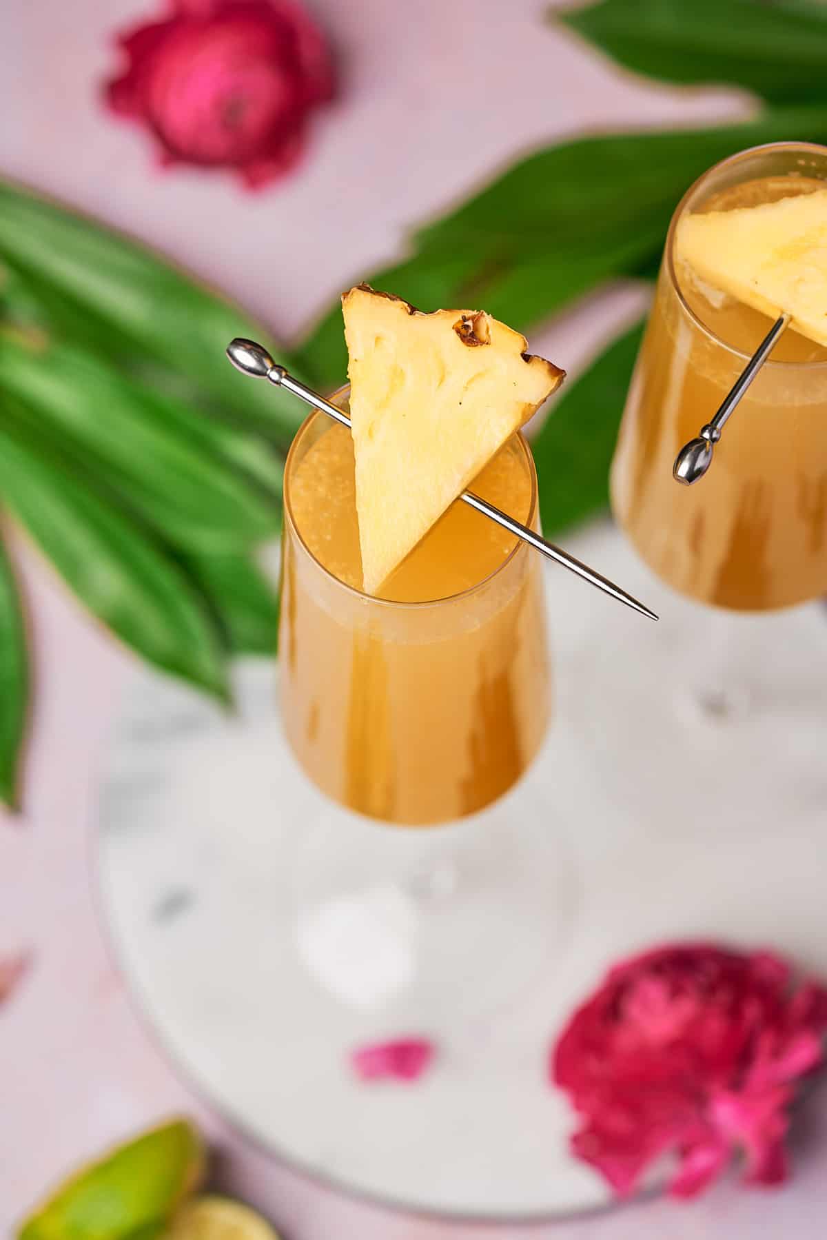 shot of two pineapple mimosas in champagne flutes, topped with a pineapple wedge, surrounded by tropical greenery and a pink florals. 