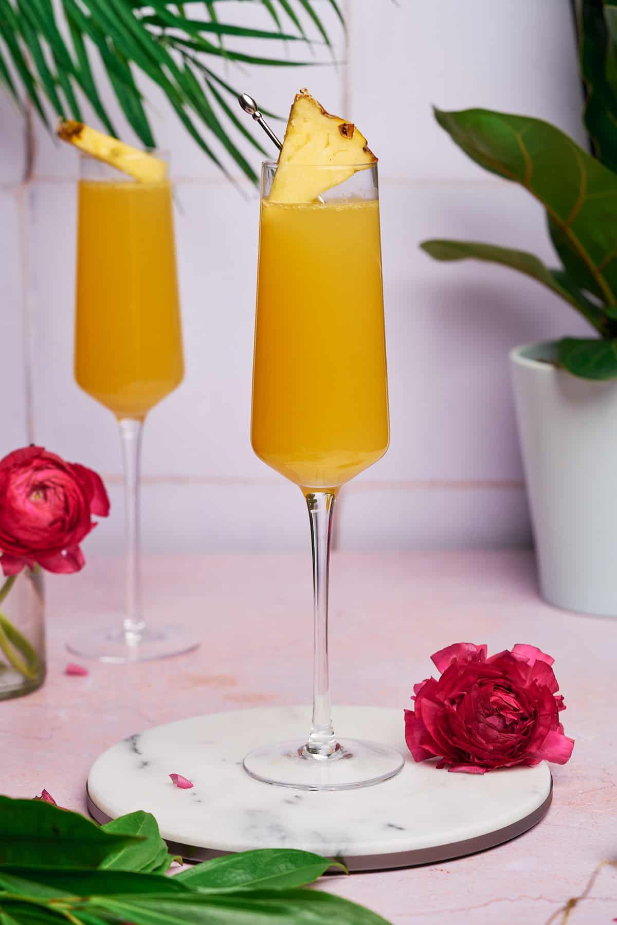 close up shot of two pineapple mimosas in champagne flutes with pink flowers surrounding them, and tropical green plants. 