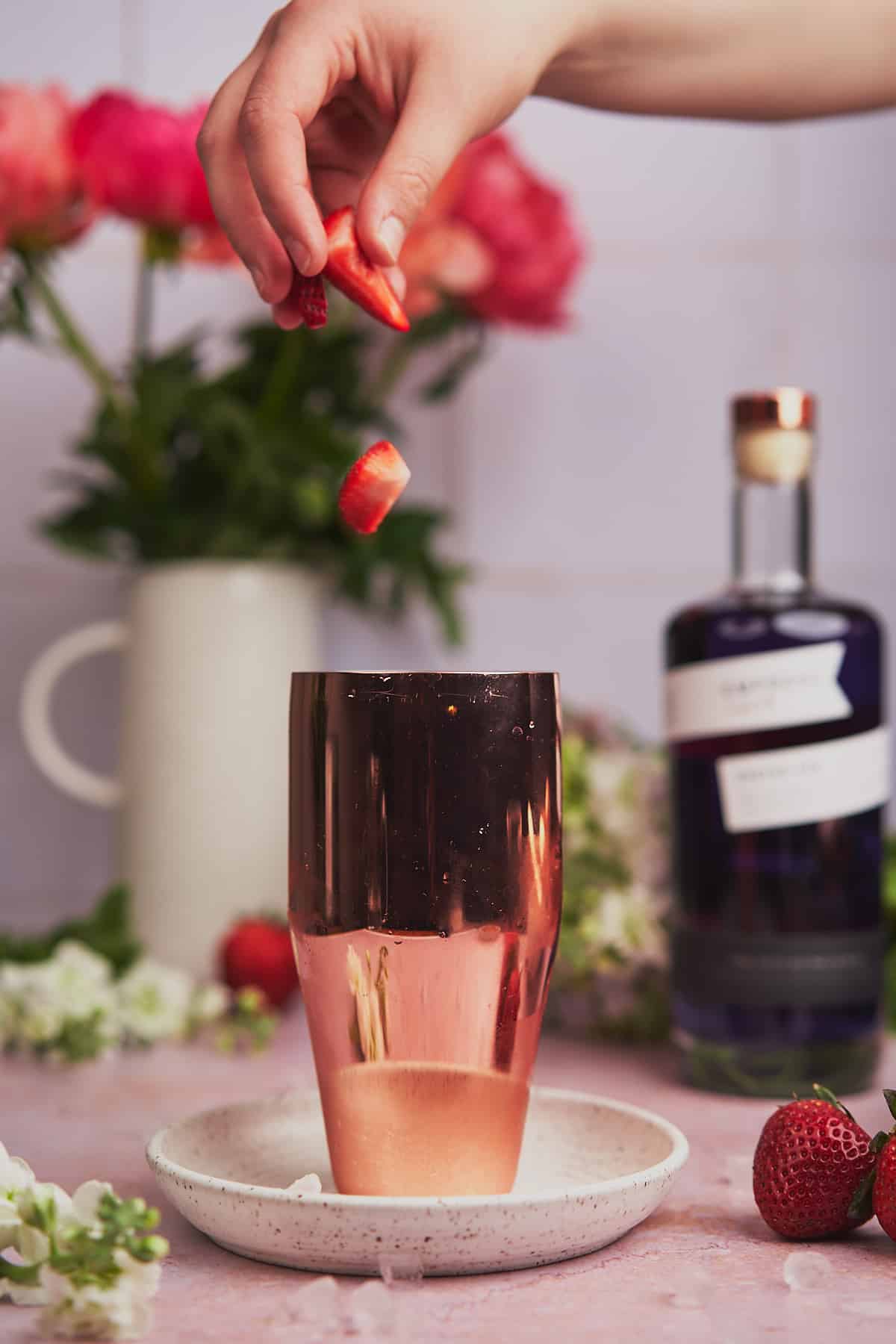 hand dropping strawberries into a cocktail shaker with empress gin and flowers in the background. 