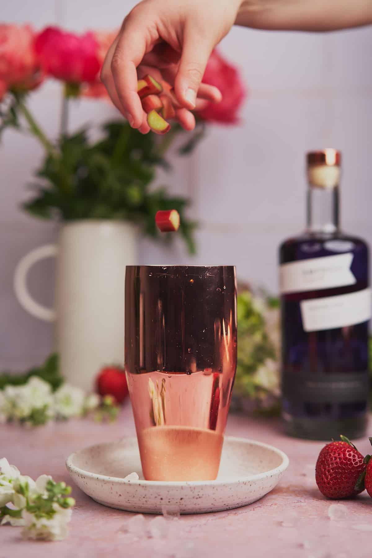 hand dropping rhubarb into a cocktail shaker with empress gin and flowers in the background. 