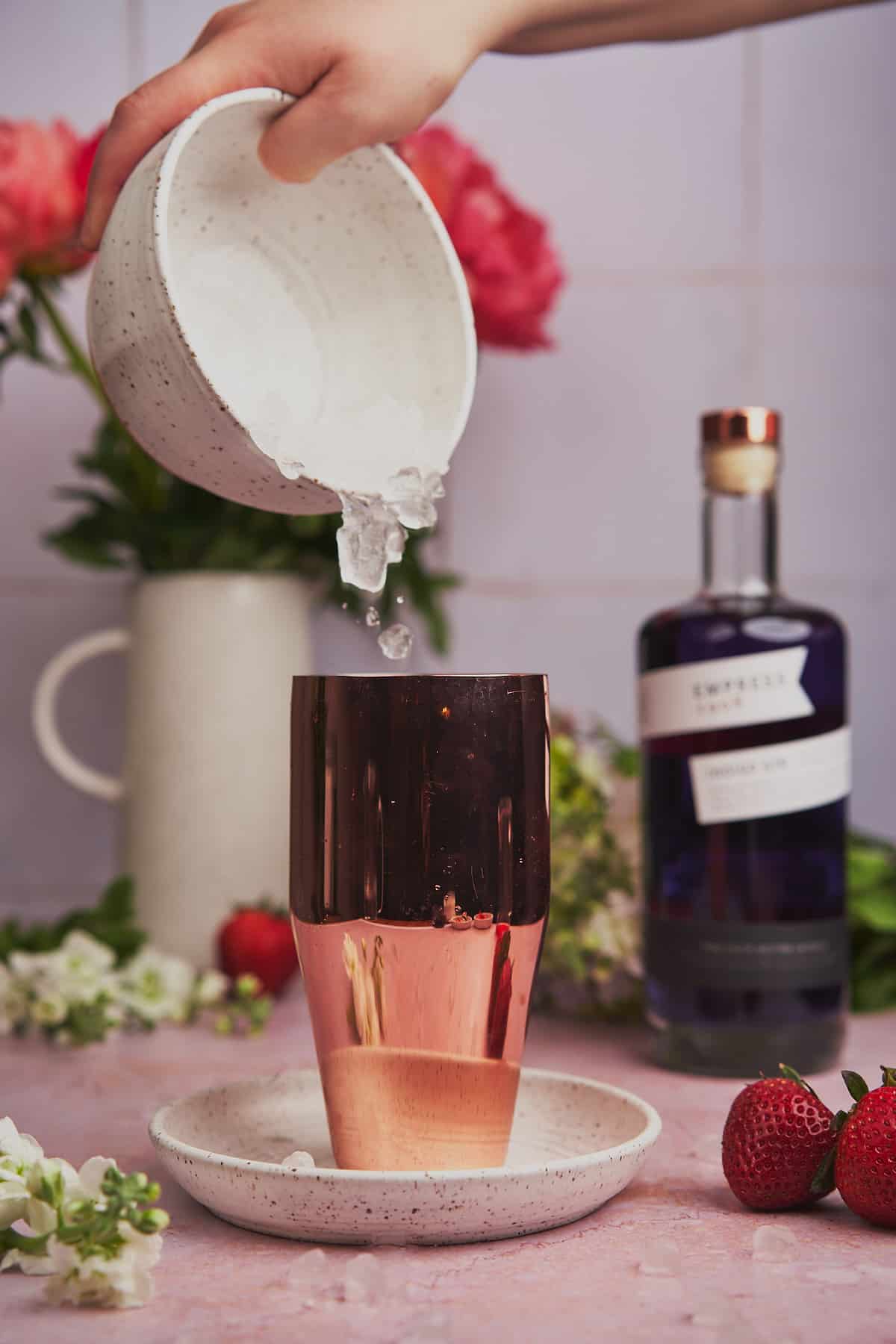 pouring ice into a cocktail shaker surrounded by fresh flowers, and Empress 1908 gin. 
