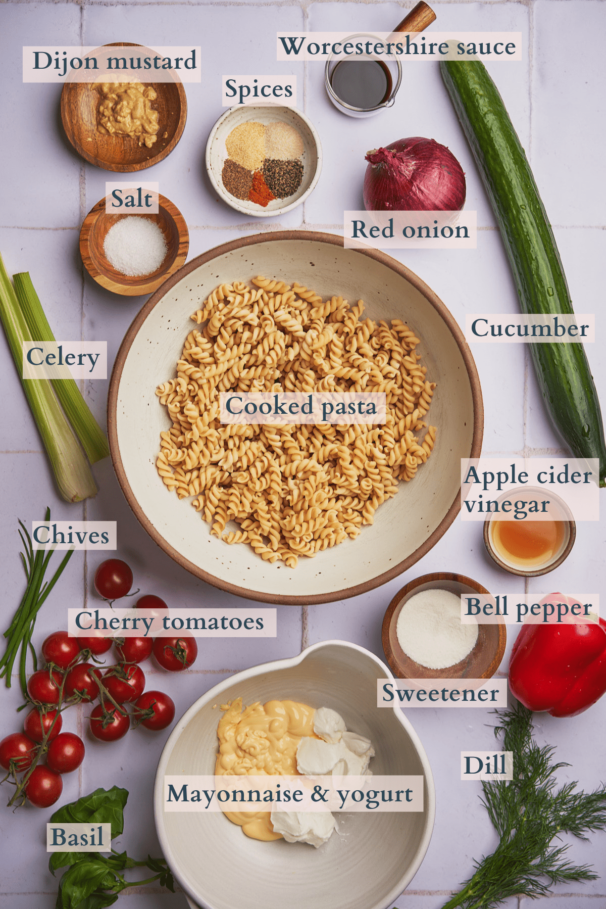 ingredients to make creamy pasta salad, with cucumbers, red onion, tomatoes, bell peppers, mayonnaise, greek yogurt, apple cider vinegar, sweetener, red bell peppers, celery, salt, spices, worcestershire sauce, dijon mustard, basil, dill, and chives.