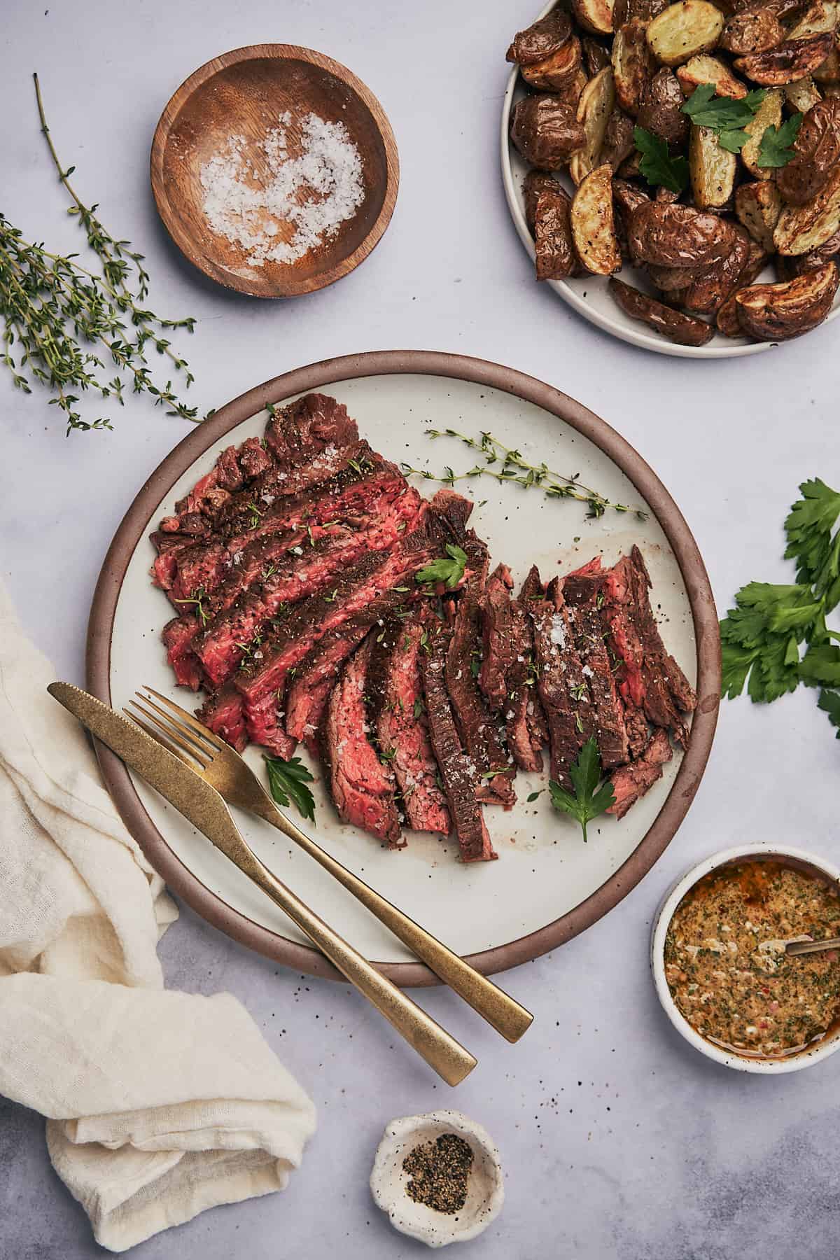 zoomed out flat lay scene of bavette steak cut up on a plate, served with flaky sea salt, pepper, parsley, thyme, buttery dipping sauce, and roasted potatoes. 