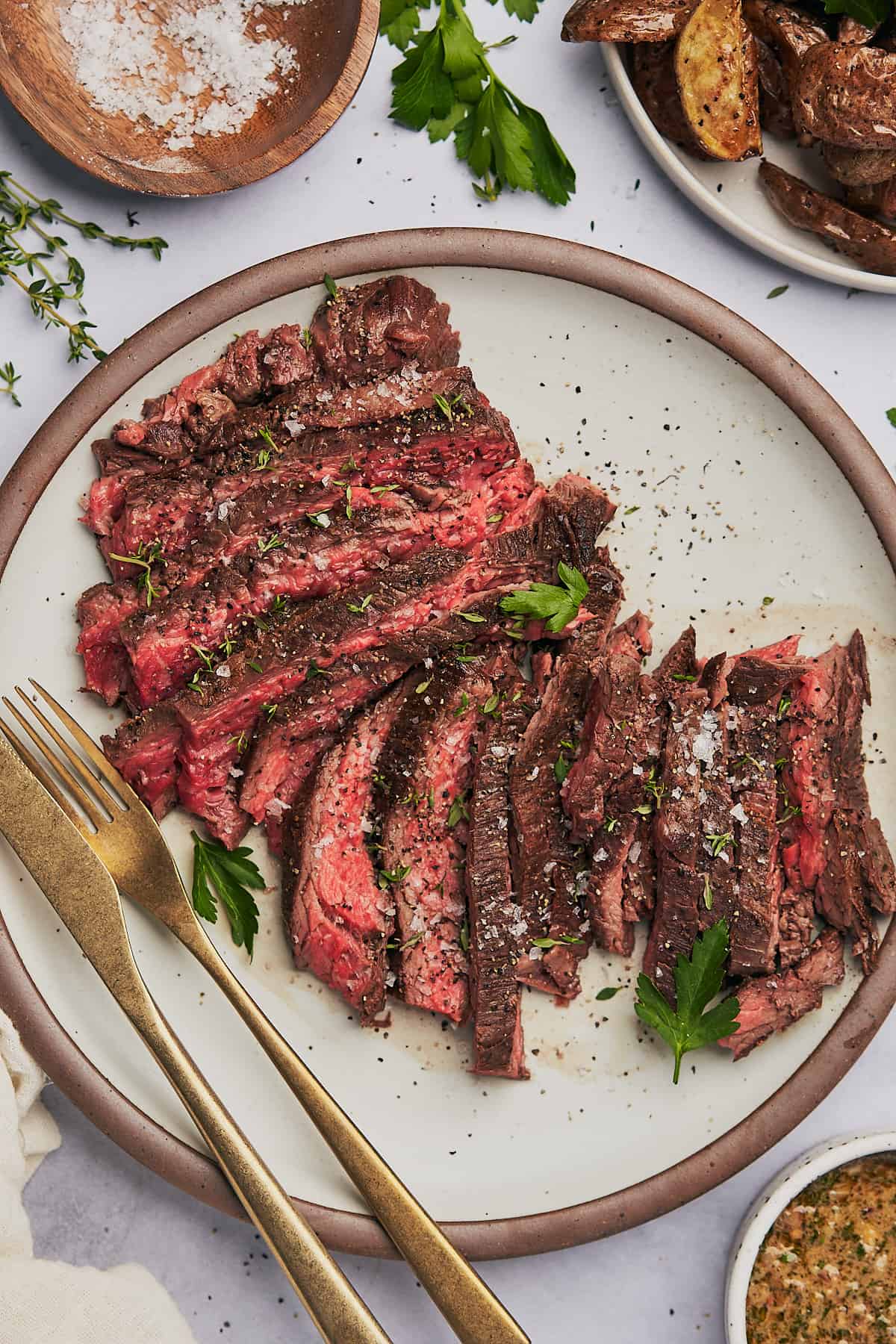 bavette steak cut nicely and served on a plate with gold silverware, parsley, thyme, potatoes, and cowboy butter dipping sauce. 