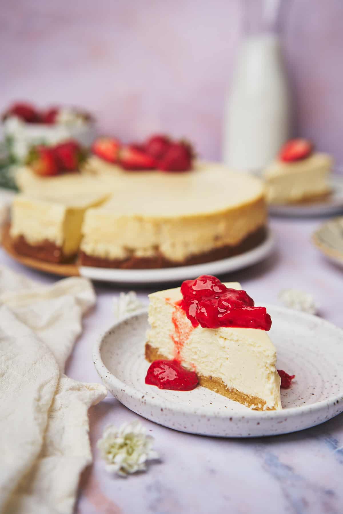 keto cheesecake recipe topped with a fresh strawberry topping, milk, and flowers in the background. 