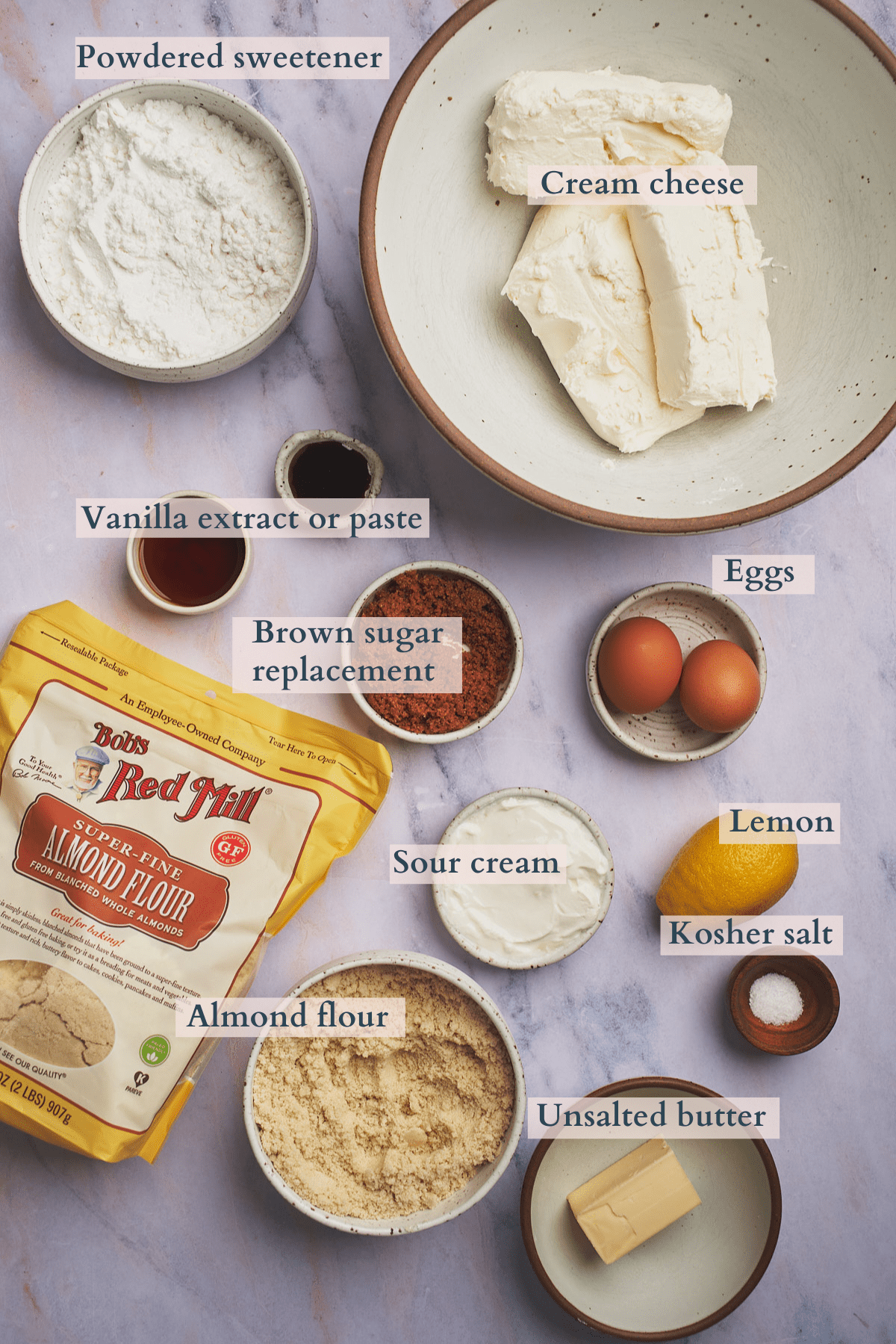 ingredients to make keto cheesecake with sour cream, cream cheese, powdered sweetener, brown sugar replacement, vanilla extract, kosher salt, eggs, lemon, butter, and almond flour.