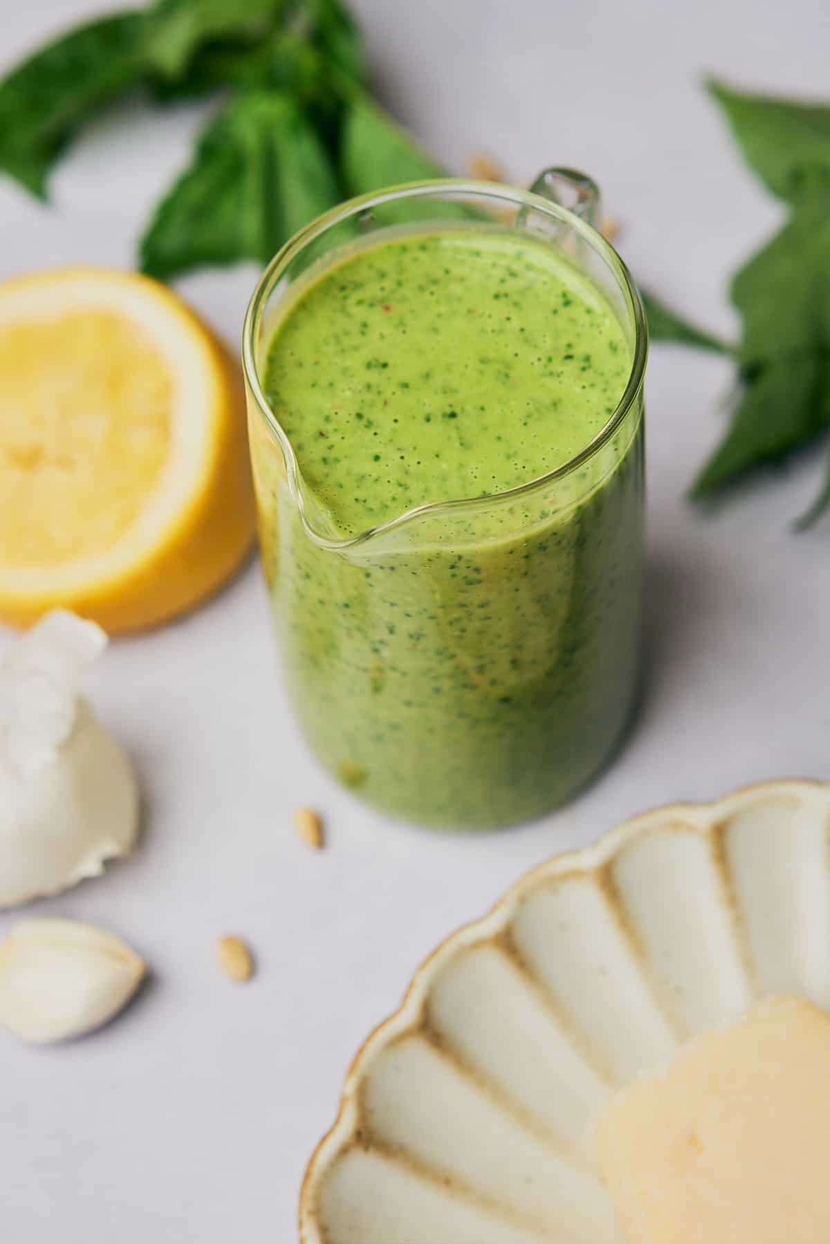 close up shot of creamy pesto vinaigrette dressing with a ruffled plate, garlic, lemon, and fresh basil nearby. 