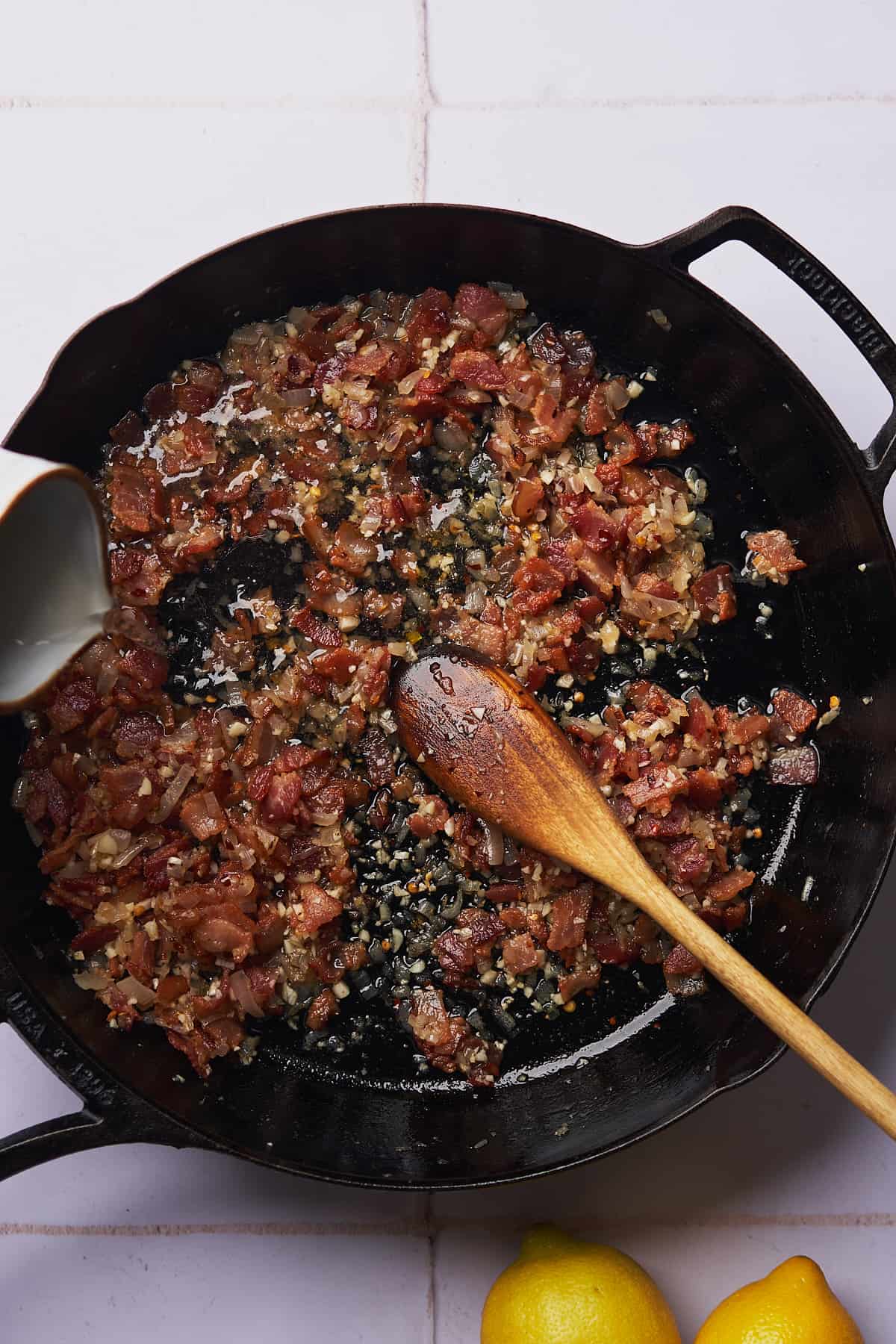 pouring white wine into a pan with bacon bits, shallots, and garlic. 