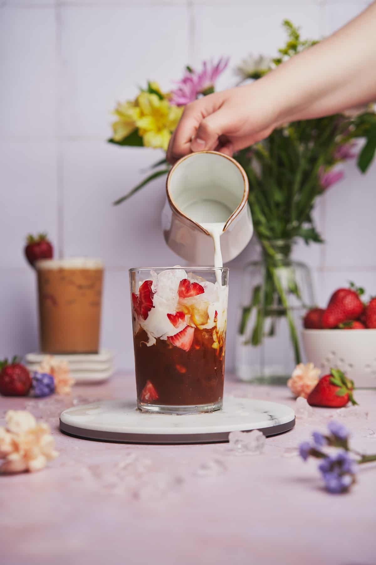 creamy strawberry latte recipe, with a hand pouring cold foam over top of a strawberry filled iced latte, and flowers and fresh strawberries in the back.