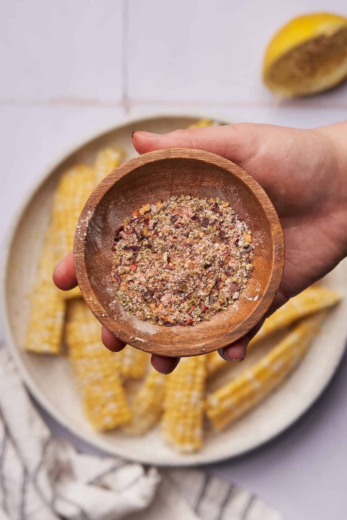 hand holding a small wooden bowl with a seasoning mixture over top of corn ribs. 