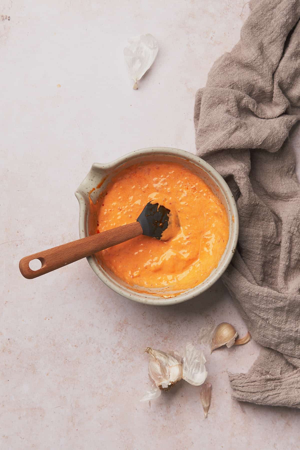 chili garlic mayo in a small gray ceramic bowl with a rubber spatula, and garlic cloves laying around it. 