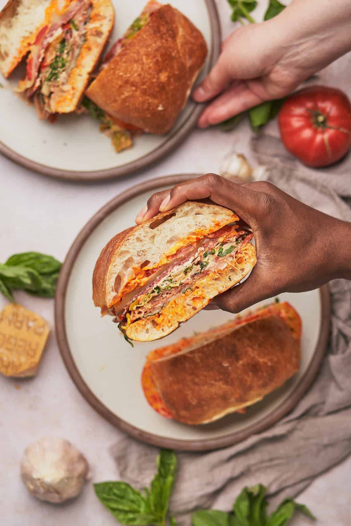 man holding an italian sub cut in half filled with meats, cheeses, veggies, and spicy mayo, with a woman reaching for her sub. 