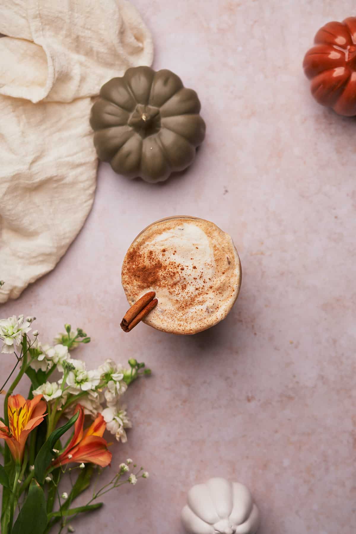 one small iced pumpkin spice latte with a cinnamon stick, and ground cinnamon over top, with flowers and ceramic pumpkins nearby. 