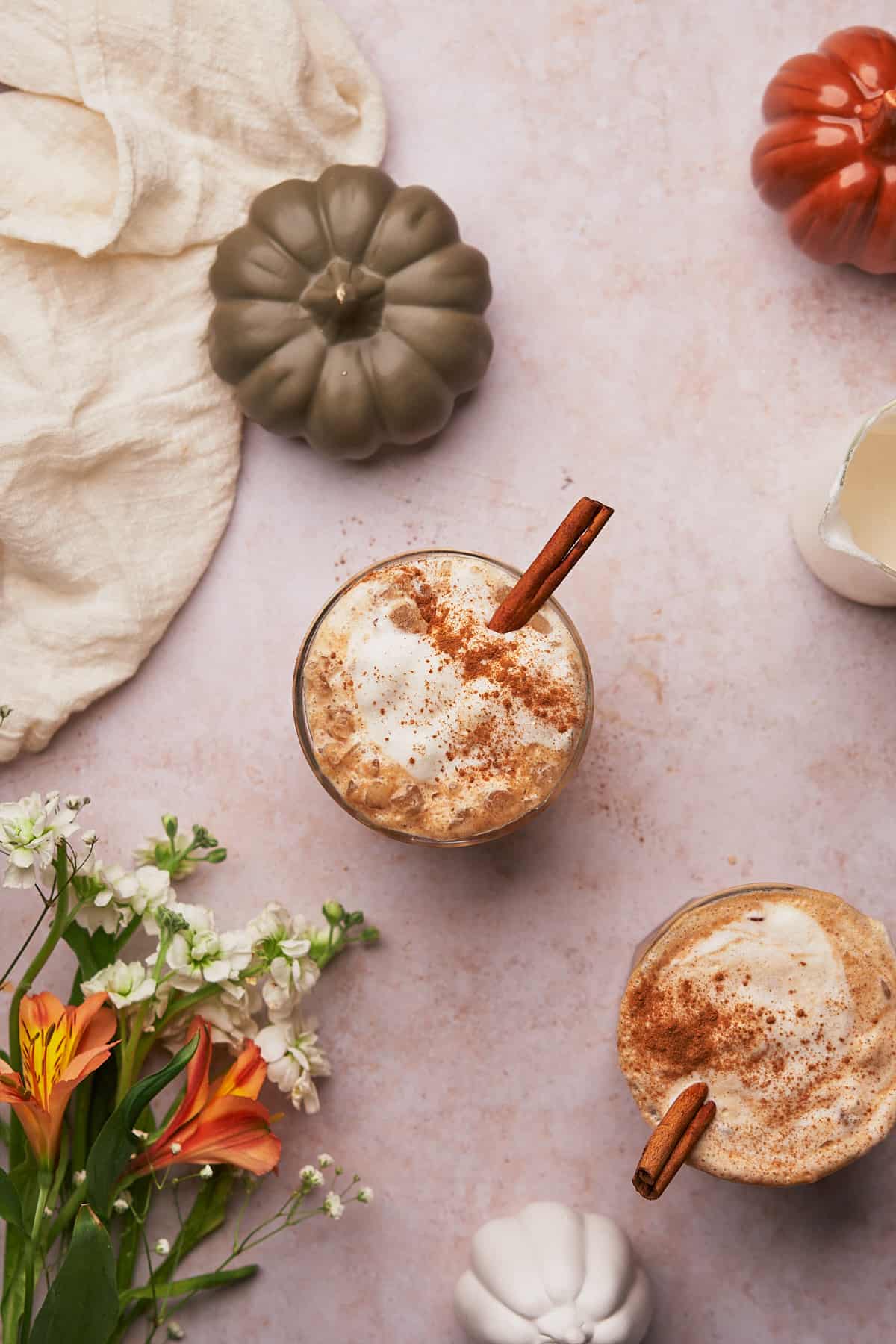 two iced pumpkin spice lattes with cinnamon sticks, and dried cinnamon sprinkled over topm surrounded. by olive green and orange small ceramic gourds and flowers.