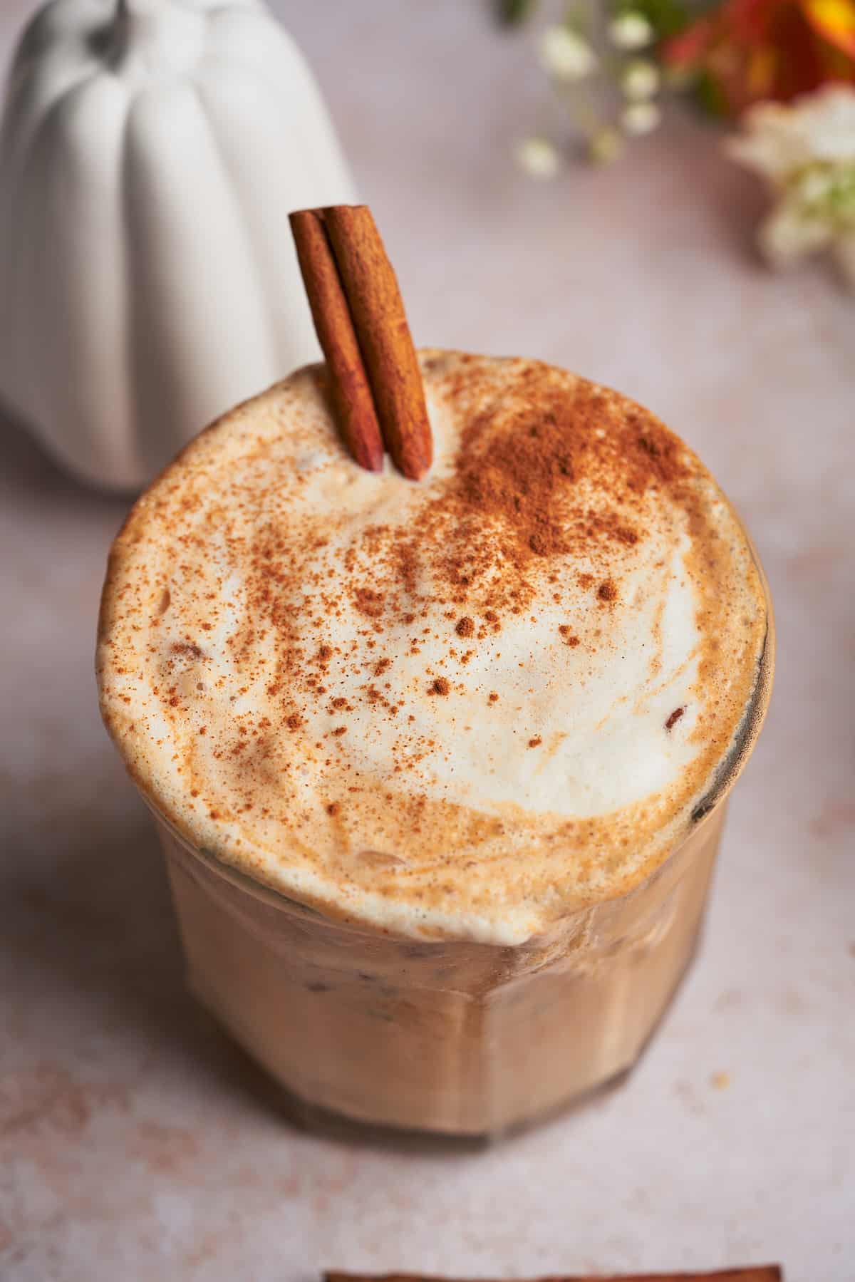 close up shot of a latte topped with ground cinnamon, heavy cream foam, and a cinnamon stick, with a ceramic pumpkin in the background. 