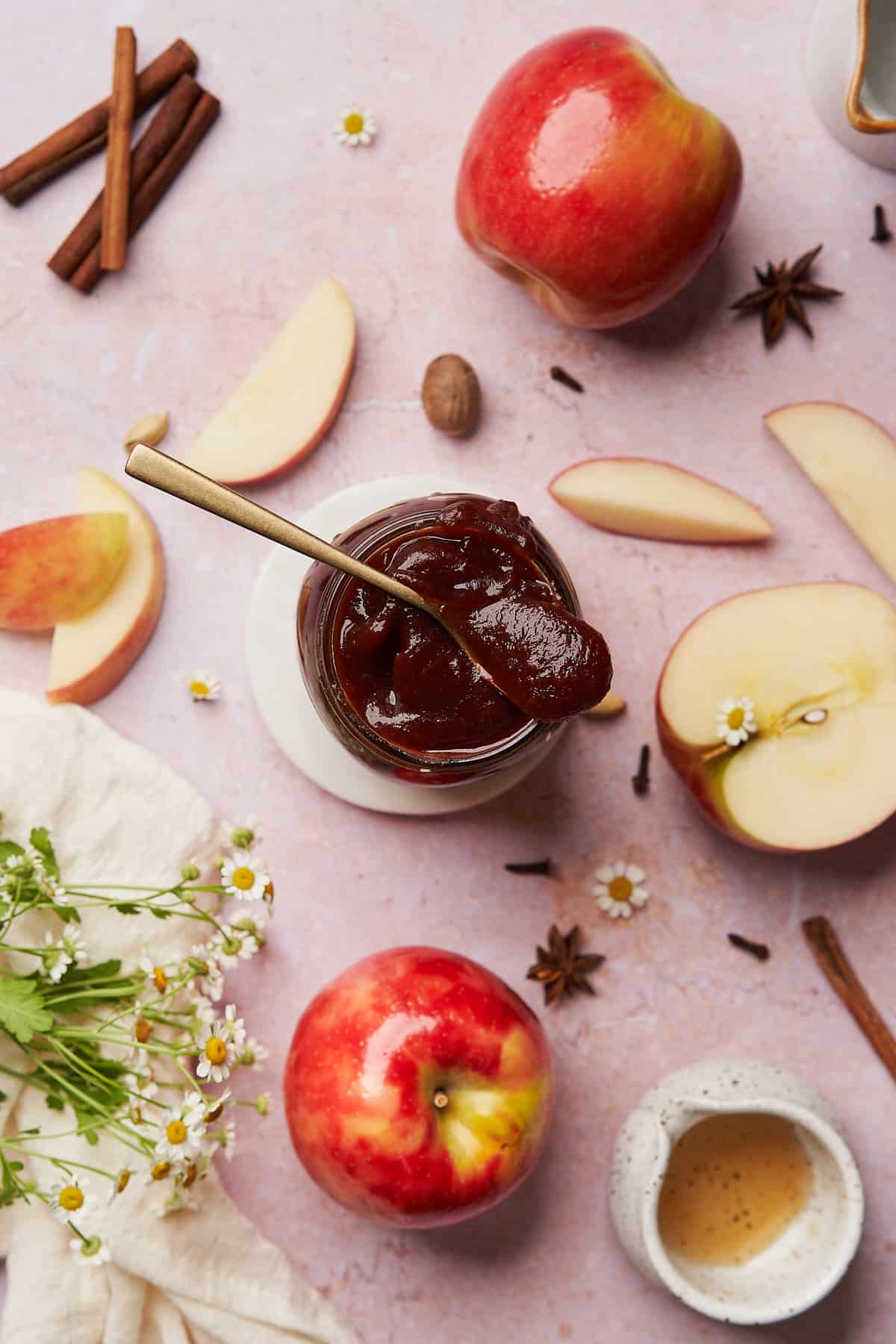 mason jar of apple butter, with a spoon over top of the jar and fresh apples and whole spices surrounding the jar. 