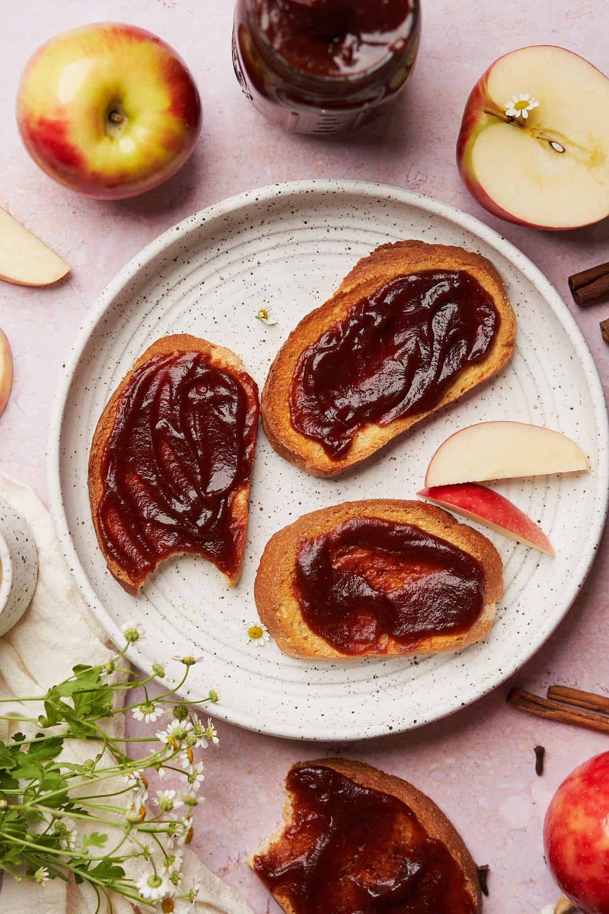 3 pieces of toast with apple butter, small chamomile flowers, cinnamon sticks and fresh apples nearby. 